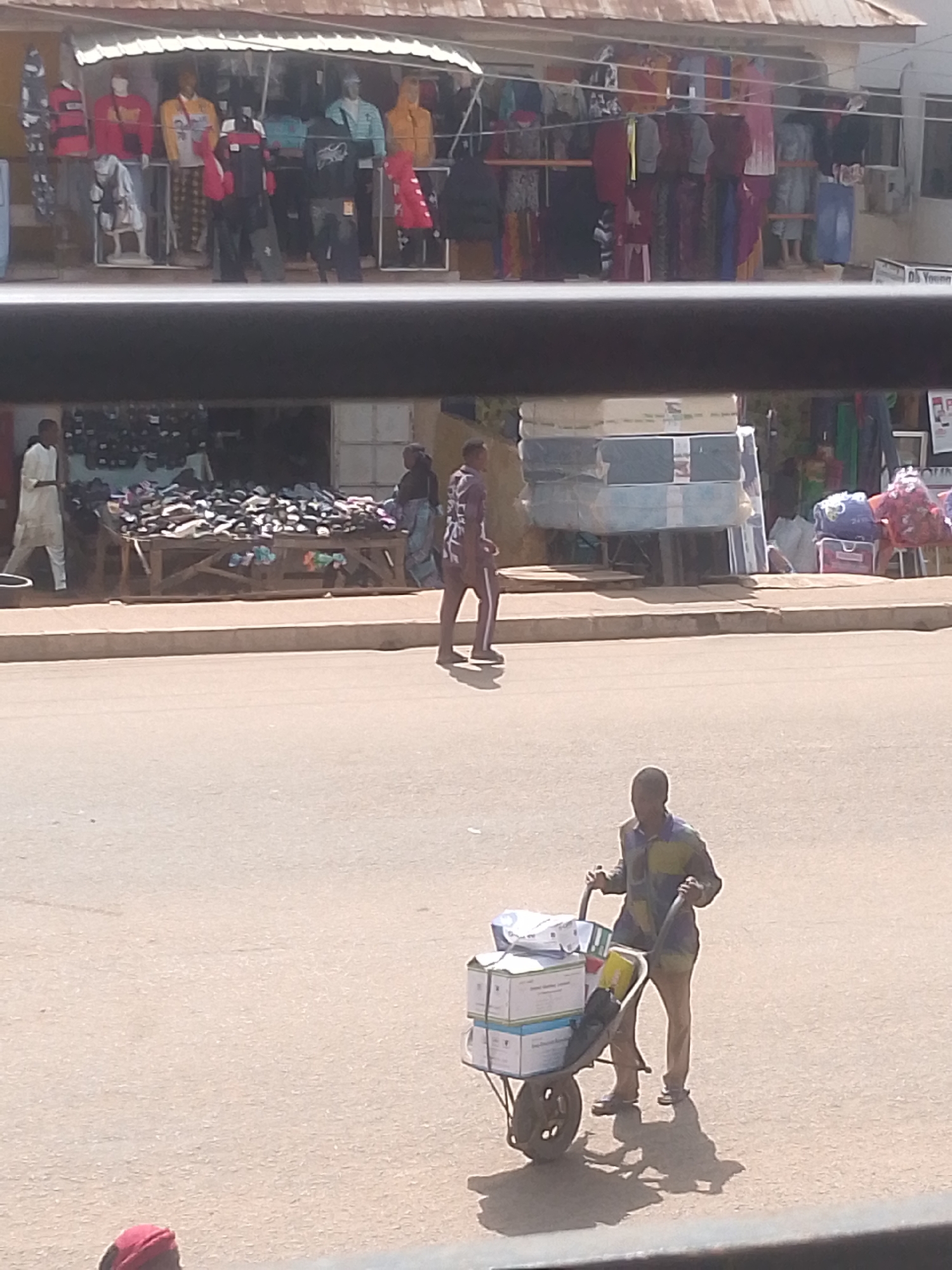 In Bukuru, Nigeria, a street scene unfolds in front of a market stall displaying clothing on mannequins and racks. A man in a white robe walks to the left, while another person in a patterned outfit walks to the right. A large table laden with shoes is visible in front of the stall. Further down the street, a man pushes a wheelbarrow filled with boxes, casting a shadow on the dusty ground. The market appears to be an outdoor setup with a covered roof. A black barrier runs across the lower part of the image, obscuring the bottom edge of the scene.