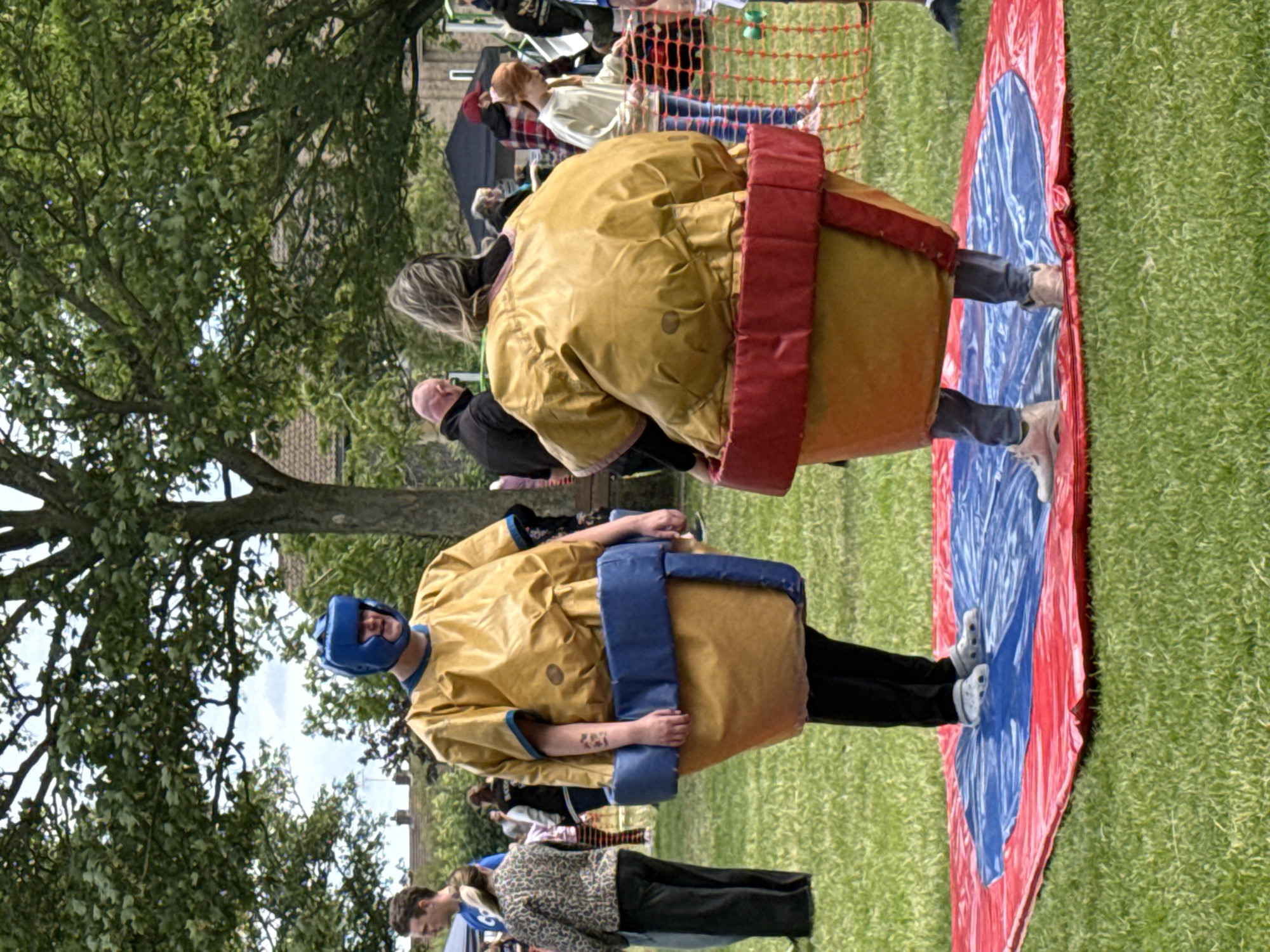 Two individuals are participating in a sumo wrestling activity on a circular inflatable mat. The mat is red on the outer edge and blue in the center, with a white swirl pattern. The participants are wearing inflatable sumo wrestling costumes that are yellow, with blue padding around the waist and red padding around the lower part of the costume. The person on the left is facing forward and wearing a blue helmet. The person on the right is facing away from the camera. Both individuals are standing on the mat. In the background, there are trees, buildings, and other people observing. The ground is covered in green grass. The scene appears to be an outdoor event, possibly a festival or fair, set in a park or open field. The location is identified as Great Yarmouth, United Kingdom.