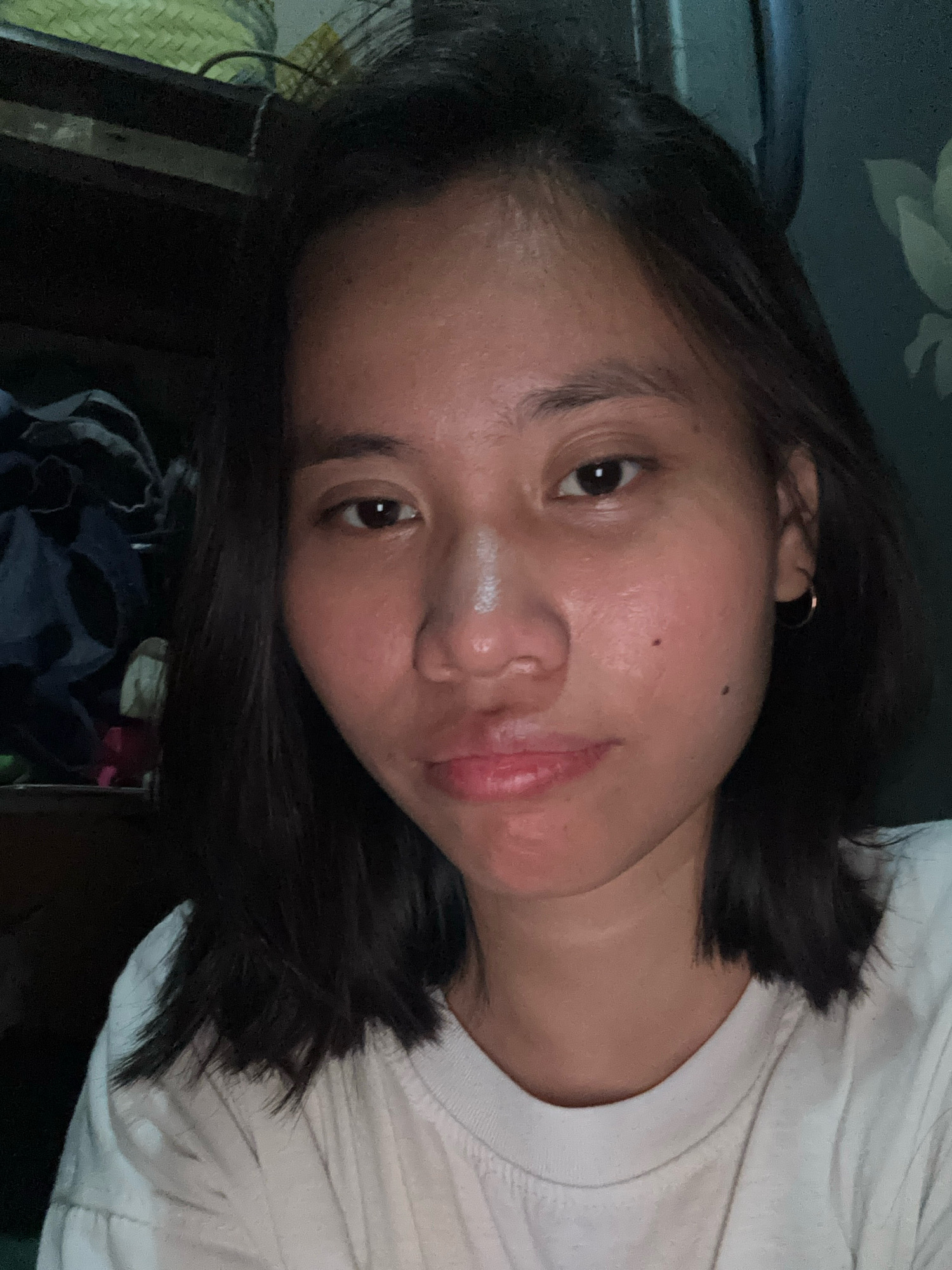 The image is a close-up selfie of a young woman with dark hair and brown eyes, wearing a white t-shirt. She has a small hoop earring in her right ear and a mole on her right cheek. The background is dimly lit and appears to be an indoor setting, with some shelves and fabric visible. The woman's face is illuminated by what appears to be artificial light, creating some shine on her skin. The image does not provide any specific details that would allow for the determination of its location in East Jakarta, Indonesia.