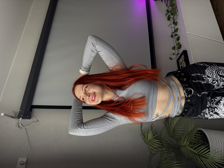 A single female subject, appearing to be in her 20s to early 30s, with long, straight, bright red hair and a septum piercing, is depicted posing indoors in a location identified as Rostov-on-Don, Russia. She is smiling and looking upwards, with both hands resting behind her head. The subject wears a light grey, long-sleeved crop top featuring straps that wrap around her midriff, revealing a tattoo on her left side, just above the hip. She is also wearing black jeans with white graphic patterns and a black belt with a silver heart-shaped buckle and eyelets. A white smartwatch is visible on her left wrist.
The environment features a light grey wall. To the upper left, a white electrical outlet and a wall-mounted white light fixture or sensor with exposed white cabling are visible. A large, grey, retractable projector screen with a black casing is mounted on the wall behind the subject. A tall, potted green plant with fronds is positioned to the subject's left. To the right, a low, white cabinet or media console holds a trailing green vine plant and a black tray containing small, dark objects. A magenta LED light strip illuminates the wall area above the trailing plant.