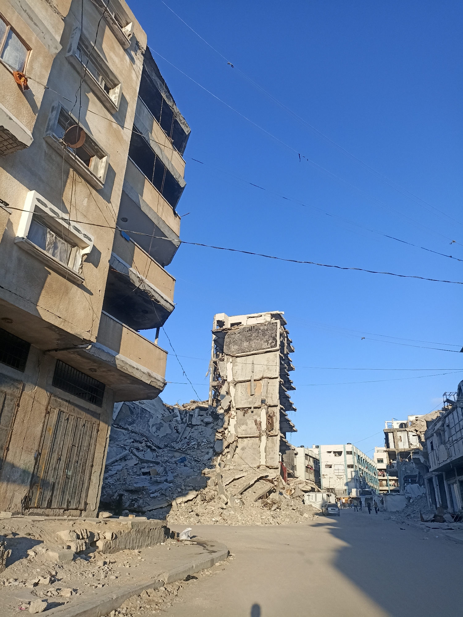 The image shows a street scene in a city that has experienced significant destruction. In the foreground, on the left, is a partially damaged multi-story building with cracked walls and missing balconies. Its lower section has metal shutters. To the right of this building is a pile of rubble, indicating structural collapse. Further down the street, another tall building stands severely damaged, with its upper floors missing and debris surrounding its base.
In the background, more damaged buildings line the street, some appearing less damaged but still showing signs of wear and tear. A few vehicles, including a silver car, are visible on the street. Several people are walking in the distance, some in small groups, suggesting some level of activity despite the devastation. The sky is clear and blue, with power lines crisscrossing overhead. The ground is a mixture of paved road and dirt, with more debris scattered around. The overall impression is one of a war-torn or disaster-stricken urban environment.