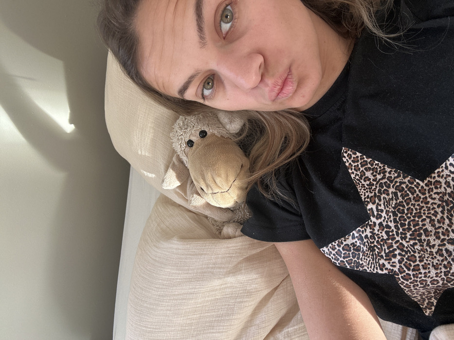 An adult female, characterized by light brown hair and light-colored eyes, is depicted in a close-up selfie. She lies on light-colored, textured bedding, wearing a black short-sleeved t-shirt with a large leopard-print star design on the front. Her lips are puckered, and she looks directly at the camera. To her left, resting on the bedding, is a light brown stuffed sheep toy with black button eyes. The scene is illuminated by natural light entering from the left, casting distinct shadows on a plain, light-colored wall in the upper background. The location is Veauchette, France.