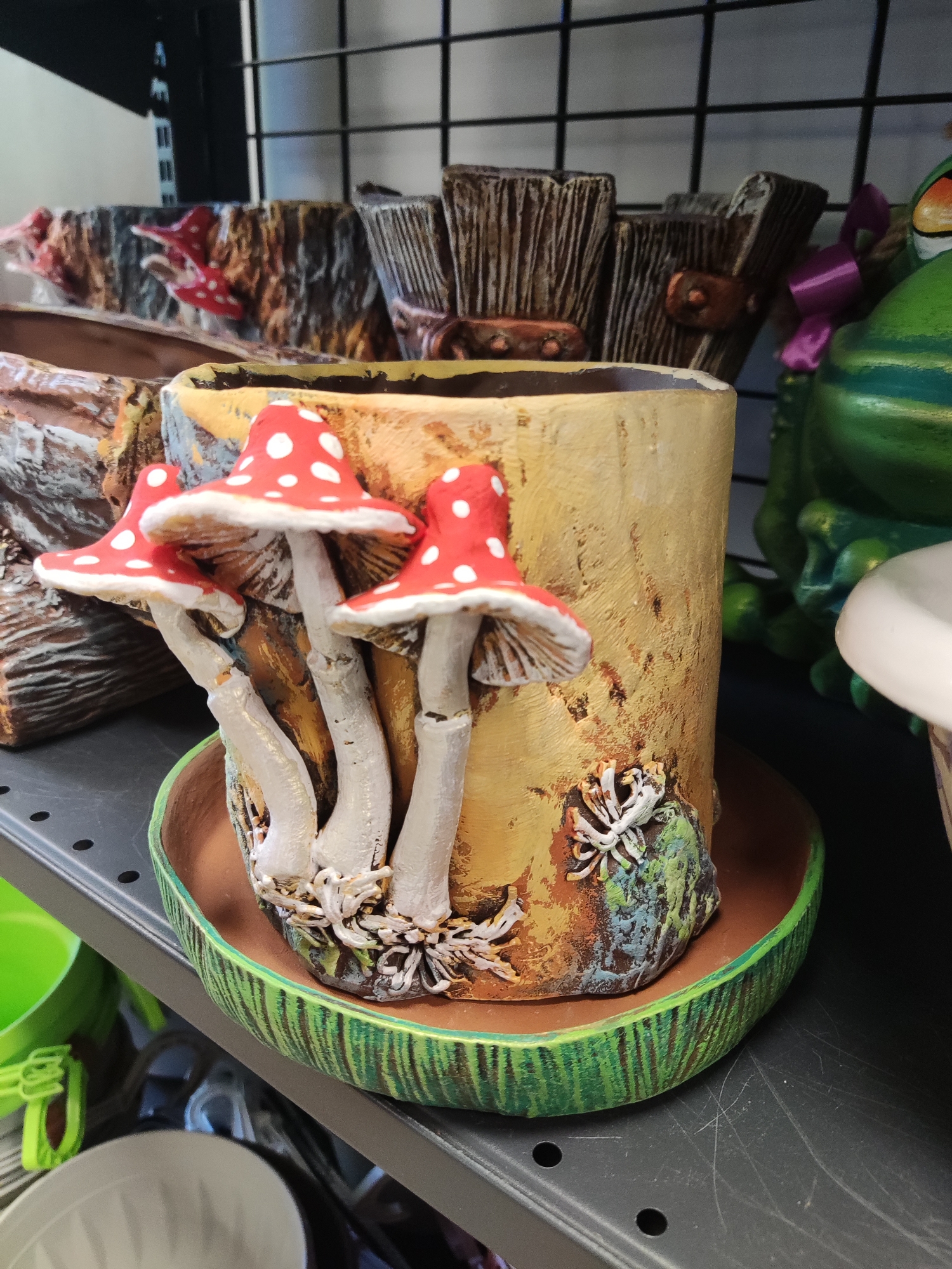 The image shows a close-up of a whimsical, mushroom-themed planter. The planter is designed to resemble a tree stump with several red and white spotted mushrooms growing from it. The mushrooms have white stems and red caps with white dots, mimicking fly agaric mushrooms. The planter itself is a cylindrical pot with a textured, wood-like exterior, painted in shades of yellow and brown. At the base of the planter, there is a small, sculpted creature that appears to be a stylized spider or insect. The planter sits on a saucer that is painted with green and brown stripes, resembling a cross-section of a log. In the background, other decorative items, including what appears to be a large green frog and more log-like planters, are visible on a dark shelf. The setting appears to be a retail environment, possibly a garden center or gift shop. The image was taken in Kryvyi Rih, Ukraine.