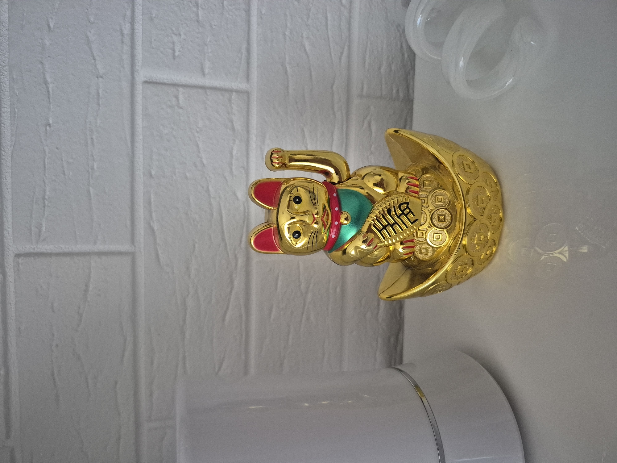 This is a close-up, eye-level, indoor photograph of a golden Maneki-neko (beckoning cat) figurine placed on a white, reflective surface. The figurine is positioned in the center of the frame, sitting on a golden boat-shaped object adorned with coin imprints. The cat has its right paw raised in a beckoning gesture and holds a golden ingot in its left. It wears a green bib and a red collar with a small golden bell. The background consists of a white brick-patterned wall. To the left of the figurine, a portion of a white cylindrical object, possibly a lamp or humidifier, is visible. To the right, two translucent white objects, appearing to be coiled tubes or rings, are present. The lighting is bright and casts soft shadows. The location of this scene is not specified as Barbing, Germany, or any other city.