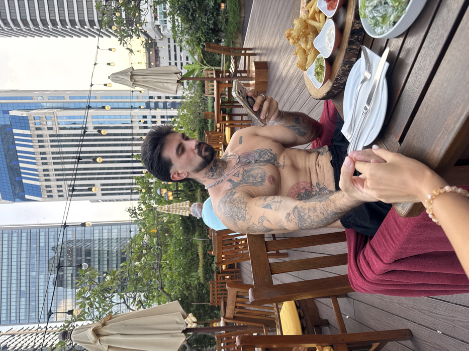 The image depicts an outdoor dining scene in a modern high-rise urban area of Bangkok, Thailand, likely an elevated terrace or rooftop restaurant.
The central figure is a male, appearing in his late 20s or early 30s, seated at a dark wooden table. He is shirtless, displaying extensive tattoos on his chest, arms, and neck, and wears black shorts. He looks down at a gold-cased smartphone held in his right hand. A silver chain with a pendant hangs around his neck, and he wears rings on his right hand. His left hand is clasped by a female's right hand, visible in the lower-left foreground, adorned with a light-colored beaded bracelet.
On the table in front of the man, a wooden serving board holds a variety of fried foods, including onion rings and french fries, alongside three small white bowls containing dipping sauces (green, white, and red). A white plate with a fork and knife is positioned next to these items. A red blanket or throw is draped over the man's chair. On the opposite side of the table, a white bowl containing a green salad is visible.
The restaurant environment features several open, light-colored patio umbrellas and string lights with exposed bulbs overhead. Lush green foliage and trees surround the dining area. In the background, multiple modern glass and concrete skyscrapers are visible under an overcast or twilight sky, with one prominent building featuring distinct blue glass sections. A blurred figure of another male in a light blue shirt is partially visible, seated at a distant table.