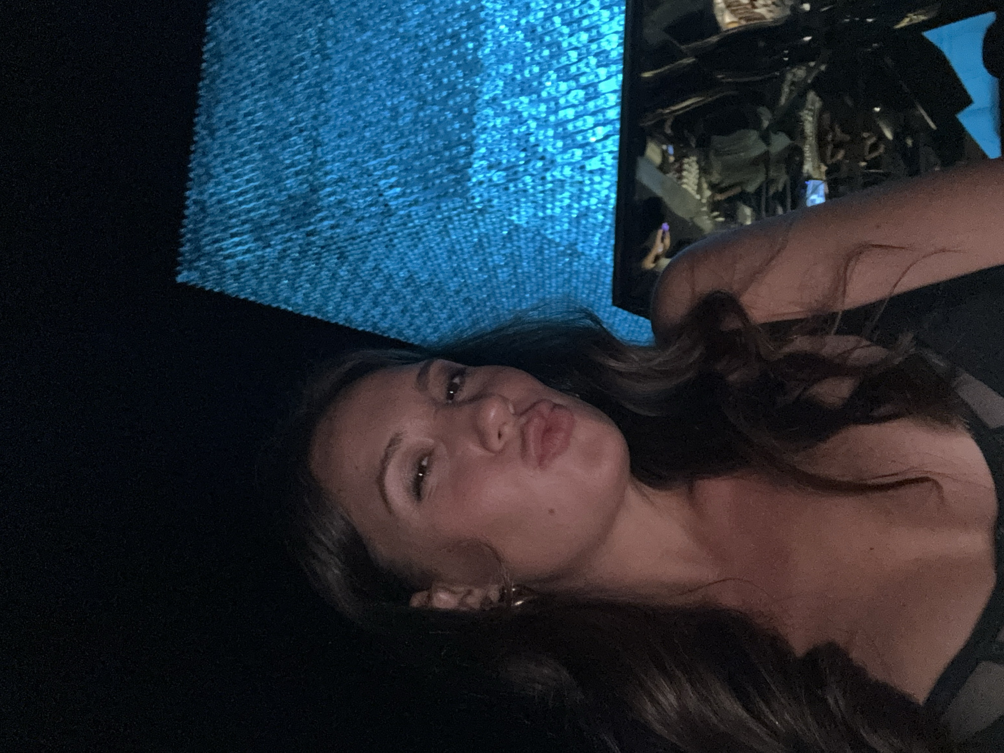 A medium close-up, horizontally oriented shot features a young woman posing playfully against a dark and abstract background. The woman, with long, dark, wavy hair cascading over her shoulders, has light-colored eyes and is making a puckered or "kissing" face directly at the camera. She appears to be wearing a dark, possibly strapless, top, with a golden hoop earring visible in her right ear.
The background is dominated by a vibrant, textured blue surface that occupies a significant portion of the upper left quadrant of the image. This blue surface appears to be composed of numerous small, reflective or illuminated elements, creating a glittering, pixelated effect, possibly a light installation or a decorative wall/ceiling. To the right, the background is darker, but reflections are visible, suggesting a mirrored or highly reflective environment. These reflections show indistinct shapes, possibly other people or architectural features, adding to the sense of a busy indoor venue.
The overall lighting is dim, with the bright blue light from the background illuminating parts of the scene and casting a cool hue. The woman's face is lit from the front, highlighting her features. The setting appears to be an indoor space, possibly a club, bar, or an artistic installation, with a modern and slightly avant-garde aesthetic. The woman's expression conveys a sense of casual fun or flirtatiousness. Based on the provided context, the location is Kuta Selatan, Indonesia. There is no visible text in the image.