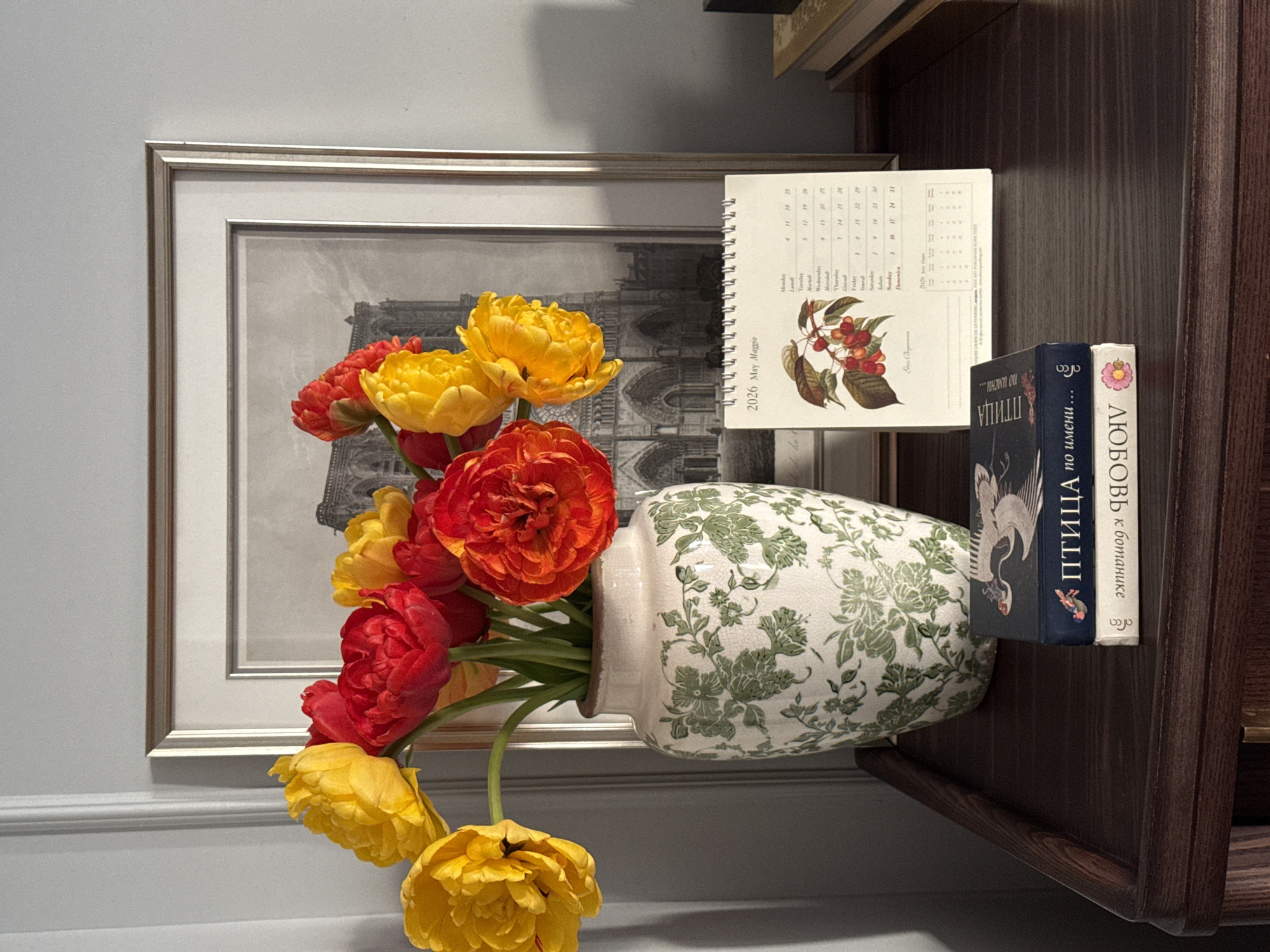 A still-life arrangement featuring a vase of colorful tulips. The flowers are predominantly red and yellow, densely packed in a white vase decorated with a green floral pattern. Behind the vase is a framed monochrome picture depicting an architectural structure with arches, possibly a cathedral. To the right of the vase, there is a calendar, open to the month of May 2026, with an illustration of cherries. Stacked next to the calendar are two books with Cyrillic text on their spines. The setting appears to be a shelf or a table against a wall, and the overall impression is of an interior home decor scene. The city of Moscow and its context in Russia are not explicitly clear in this image.