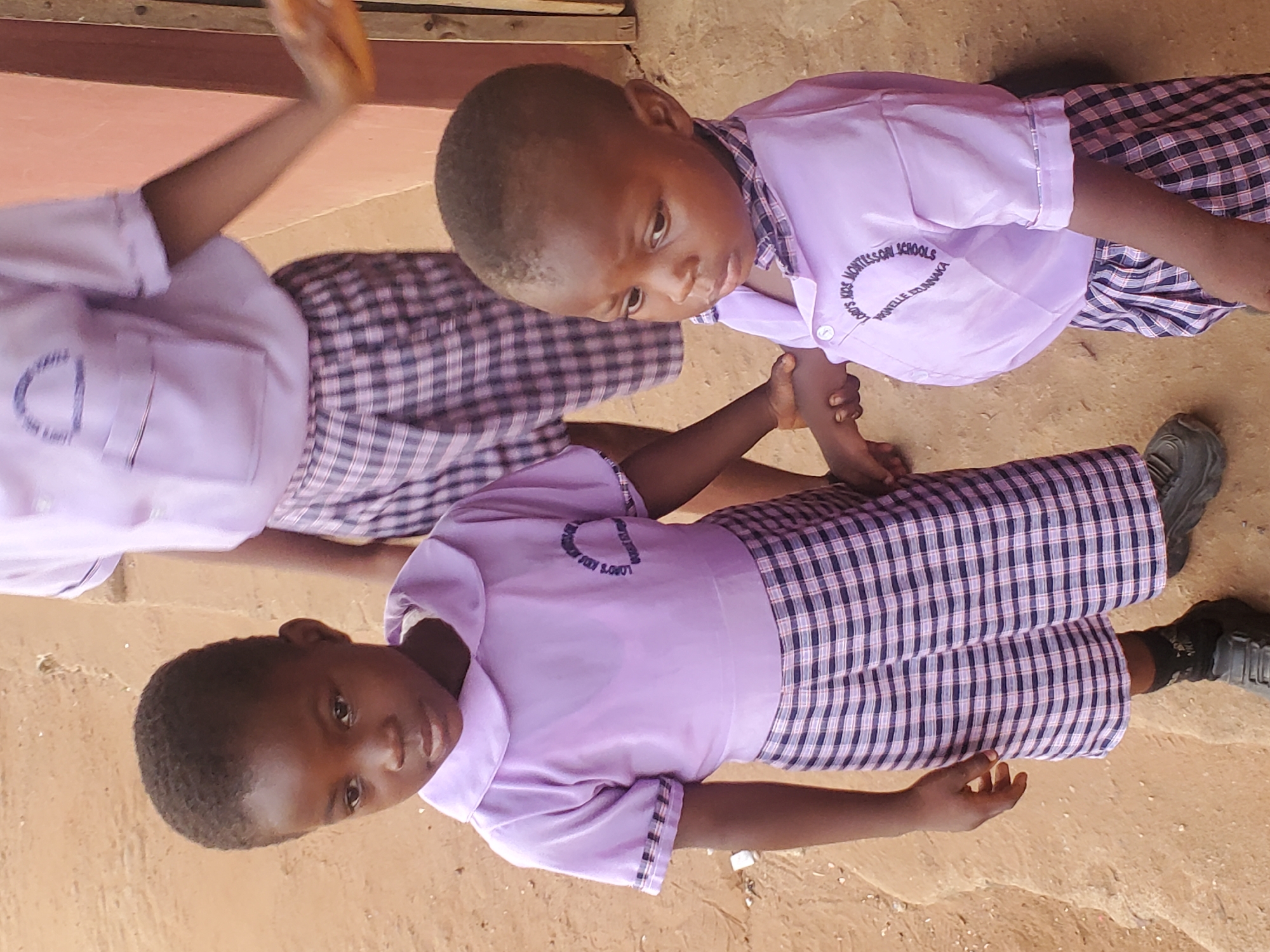 Two young children wearing school uniforms are laying on the ground, possibly outdoors in Ukwu-Abwa, Nigeria. The uniforms consist of lavender shirts with school logos and checkered pants. One child is lying with their head towards the left, while the other is oriented in the opposite direction. Both children are wearing dark shoes. The ground appears to be a dirt surface. One child is holding the wrist of another child.