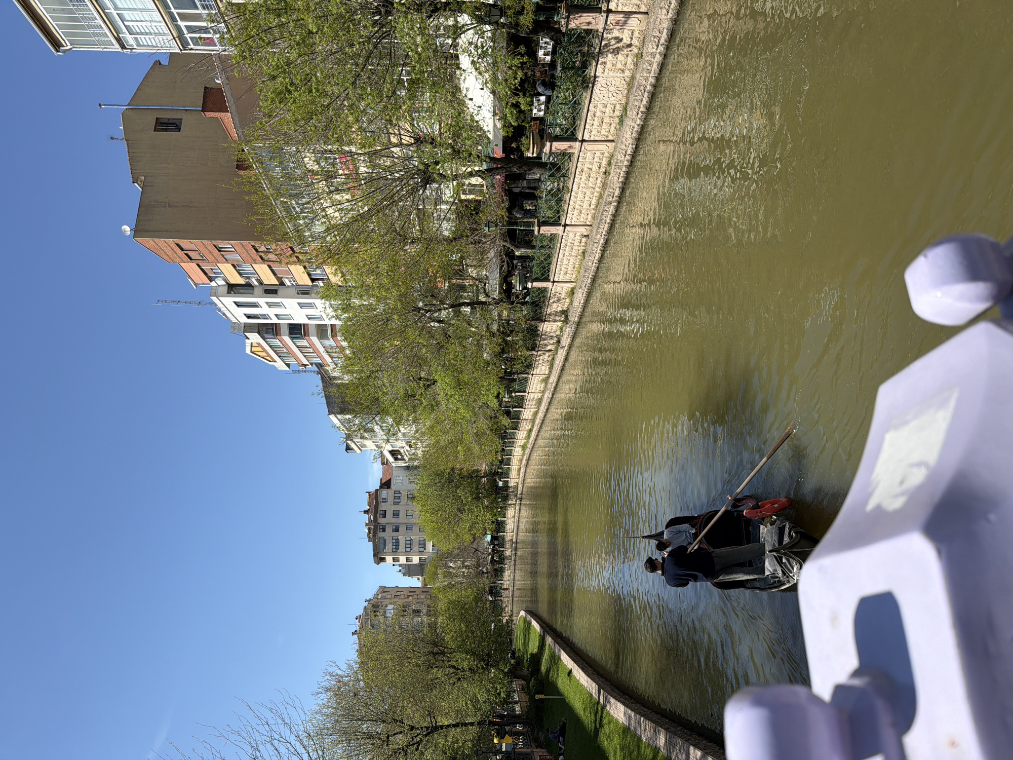 The image captures an urban canal scene in Eskisehir, Turkey, under a clear, bright blue sky. The canal, filled with green-brown water, stretches from the foreground into the distance.
In the middle of the canal, a dark-colored boat, resembling a gondola, is being actively poled by one person, while another person is seated as a passenger. A red life preserver or buoy is visible inside the boat. Ripples on the water surface reflect the daylight.
The right bank of the canal features a stone retaining wall topped with an ornate green fence. Behind this fence is a green park area lined with trees, some displaying fresh spring foliage. Interspersed among the trees are outdoor seating arrangements, including tables and chairs, suggesting cafes or restaurants alongside the canal.
On the left side of the canal, a series of multi-story buildings stand, showcasing varying architectural styles and colors, including beige, red brick, and white facades. Some buildings feature balconies, and several rooftops are adorned with satellite dishes and antennas.
The photograph appears to be taken from an elevated position, likely a bridge, with a light purple railing partially visible in the immediate foreground. The clear blue sky and strong light indicate daytime, likely morning or afternoon, with sunny weather. The overall scene depicts a calm and recreational urban environment. No discernible text is visible within the image.