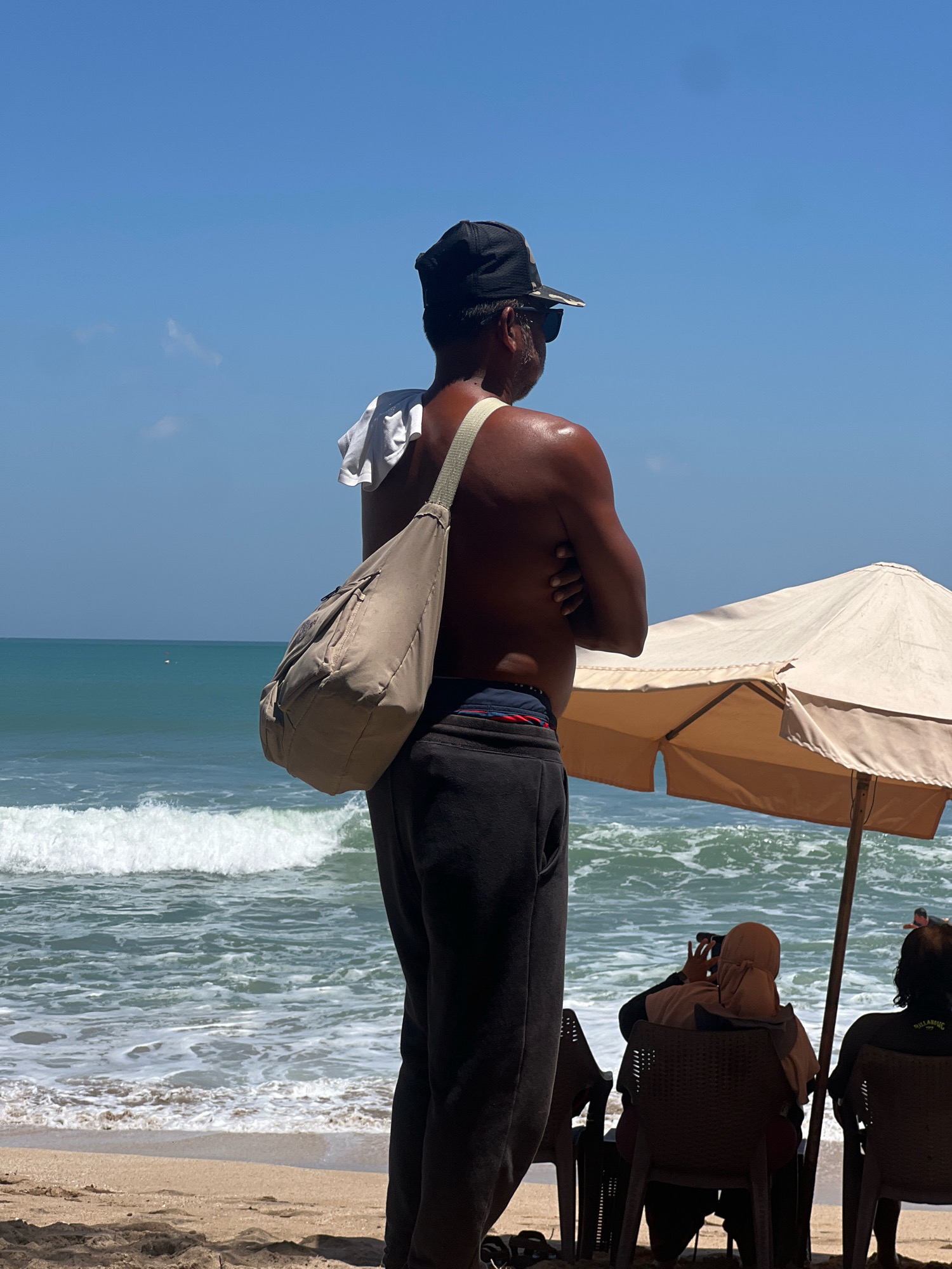 An adult male, wearing a black cap, sunglasses, and black pants, stands on the beach in Kuta, Indonesia, with his arms crossed and a light brown bag slung over his shoulder. He is facing away from the camera, looking out at the ocean. The ocean is a vibrant blue-green with white waves crashing on the shore. In the background, a beige beach umbrella provides shade for two people sitting in chairs. The sand is a light brown, with some shadows cast from the umbrella and other unseen objects. The sky is a clear, bright blue with a few wispy clouds. The scene is bathed in bright sunlight, suggesting it is midday.