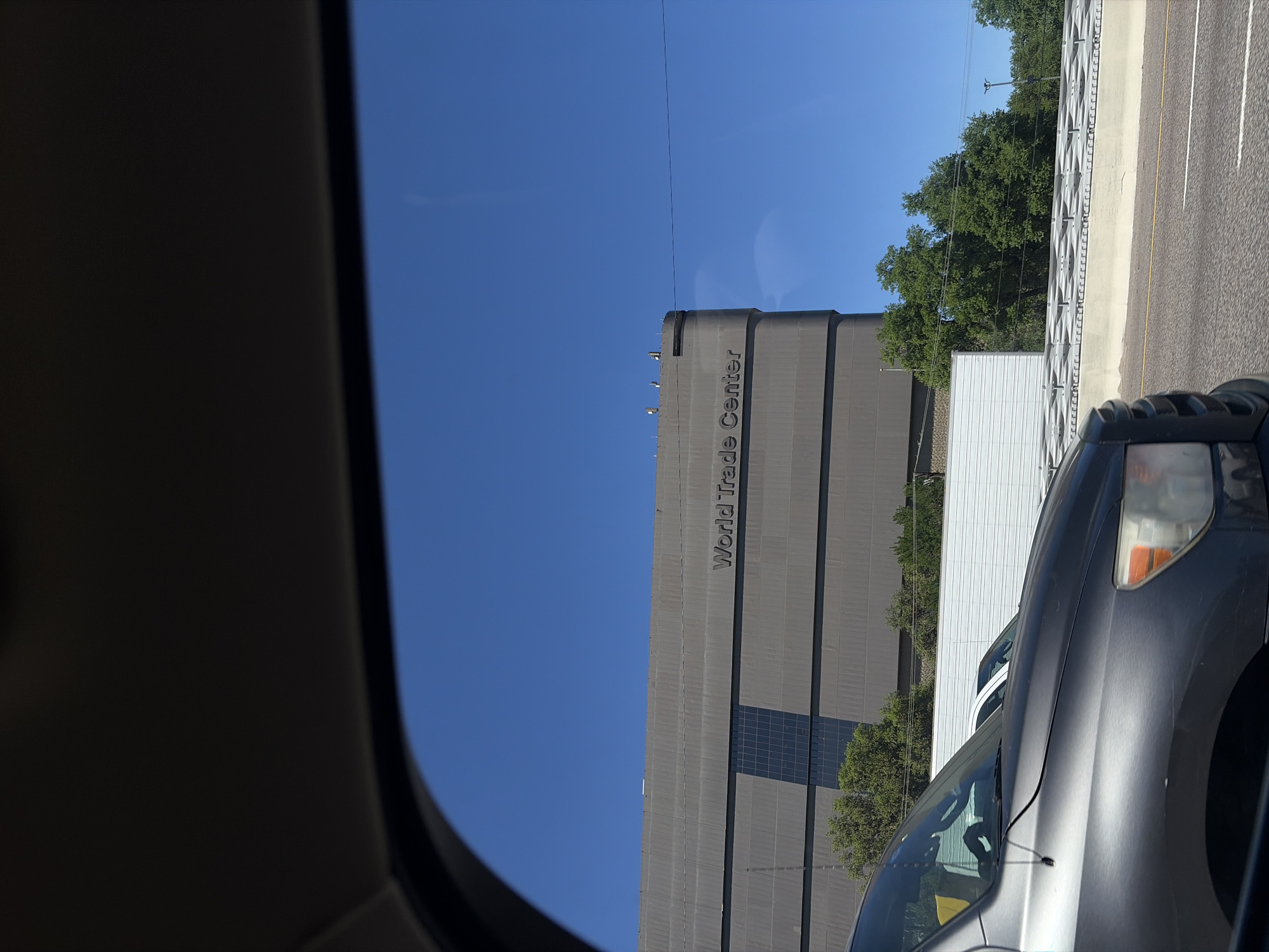 The image shows the World Trade Center in Dallas, United States of America, as viewed from inside a vehicle. The building is a tall, gray structure with the words "World Trade Center" written vertically on its side. There is a highway barrier next to it, and some trees are visible. The sky is clear and blue. Part of the car's dashboard and A-pillar are visible in the foreground of the image. Another car is in the adjacent lane.