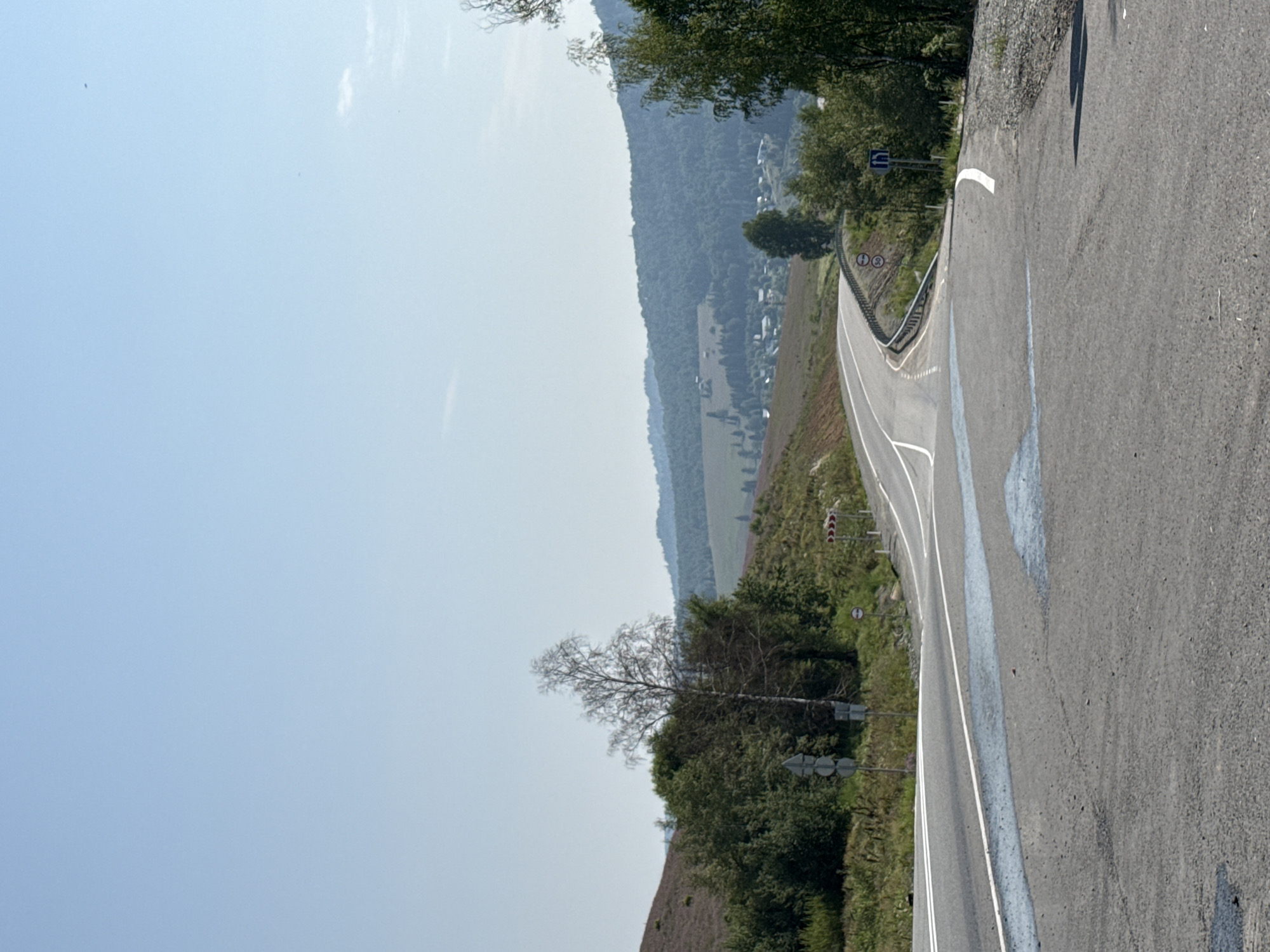 The image depicts a winding asphalt road leading through a hilly, rural landscape. The road has white lane markings and is flanked by a metal guardrail on the right side. Several traffic signs are visible, including a speed limit sign showing "30" and a sign with an upward-pointing arrow indicating a lane direction. The terrain is covered with vegetation, including trees and low-lying bushes. In the distance, there are rolling hills covered in forests, with a small village or cluster of houses visible nestled in a valley. The sky is clear and blue. The scene appears to be the outskirts of Choya, Russia, given the context of the inquiry. The image is a static, outdoor shot under daylight conditions. There are no people or visible actions occurring in the scene.
