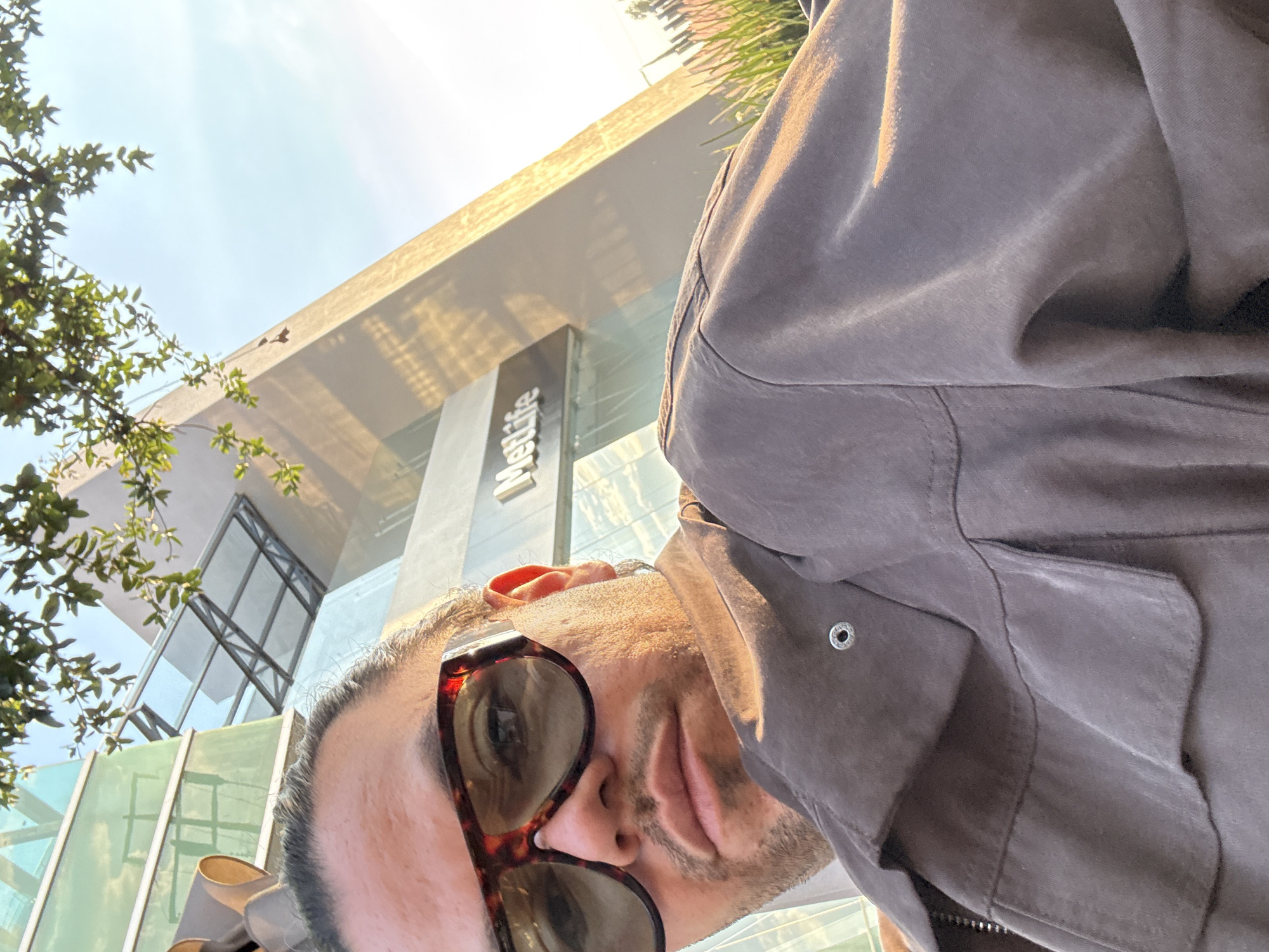 A selfie, taken in Puebla, Mexico. A man wearing tortoise shell sunglasses and a brown jacket. In the background is a tall building with glass windows. Part of the name of the building, "Angelopolis" is visible. Trees are also visible on the left side. The sun is shining in the sky.
