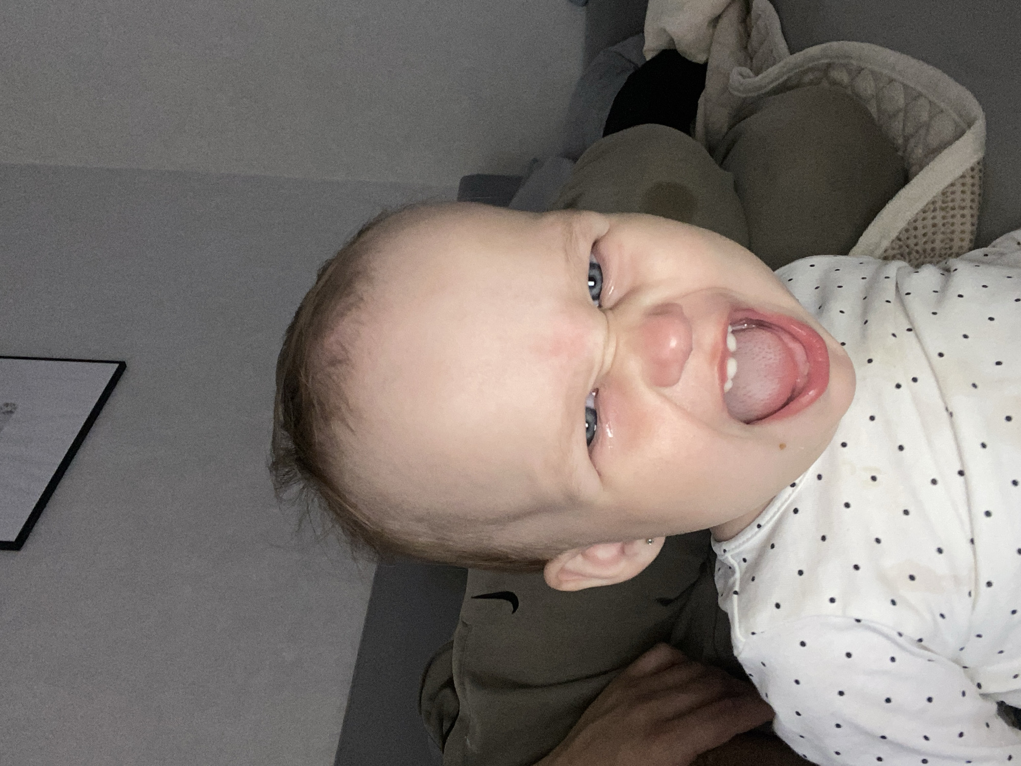 An infant, appearing to be under two years of age, is centrally positioned in a close-up, dimly lit indoor photograph. The scene is situated in the city of Chaikovsky, Russia. The infant has fair skin, sparse light brown hair, and striking blue eyes, and is captured mid-expression, with a wide-open mouth, scrunched face, and furrowed brow, consistent with a yell or intense cry. A tear is visible in the infant's left eye. The nose is wrinkled, and the mouth reveals visible lower teeth and upper gums, with the tongue partially exposed. A small, dark mole is present below the right corner of the infant's mouth. The infant is wearing a white long-sleeved garment adorned with small black polka dots, exhibiting minor brownish stains on the right shoulder and chest area. A small silver stud earring is visible in the infant's left ear. In the immediate background to the left, a portion of an adult's hand with light skin tone is visible. Directly behind the infant is a soft, muted olive green or grey furnishing, possibly a large pillow or a sofa back, featuring a distinct black Nike swoosh logo. To the infant's right, a textured light grey material, potentially a patterned blanket or cushion, is present. The walls are a plain light grey, appearing textured due to the image's graininess. A segment of a black-framed picture is visible on the upper left wall. The ambient lighting is low and artificial.