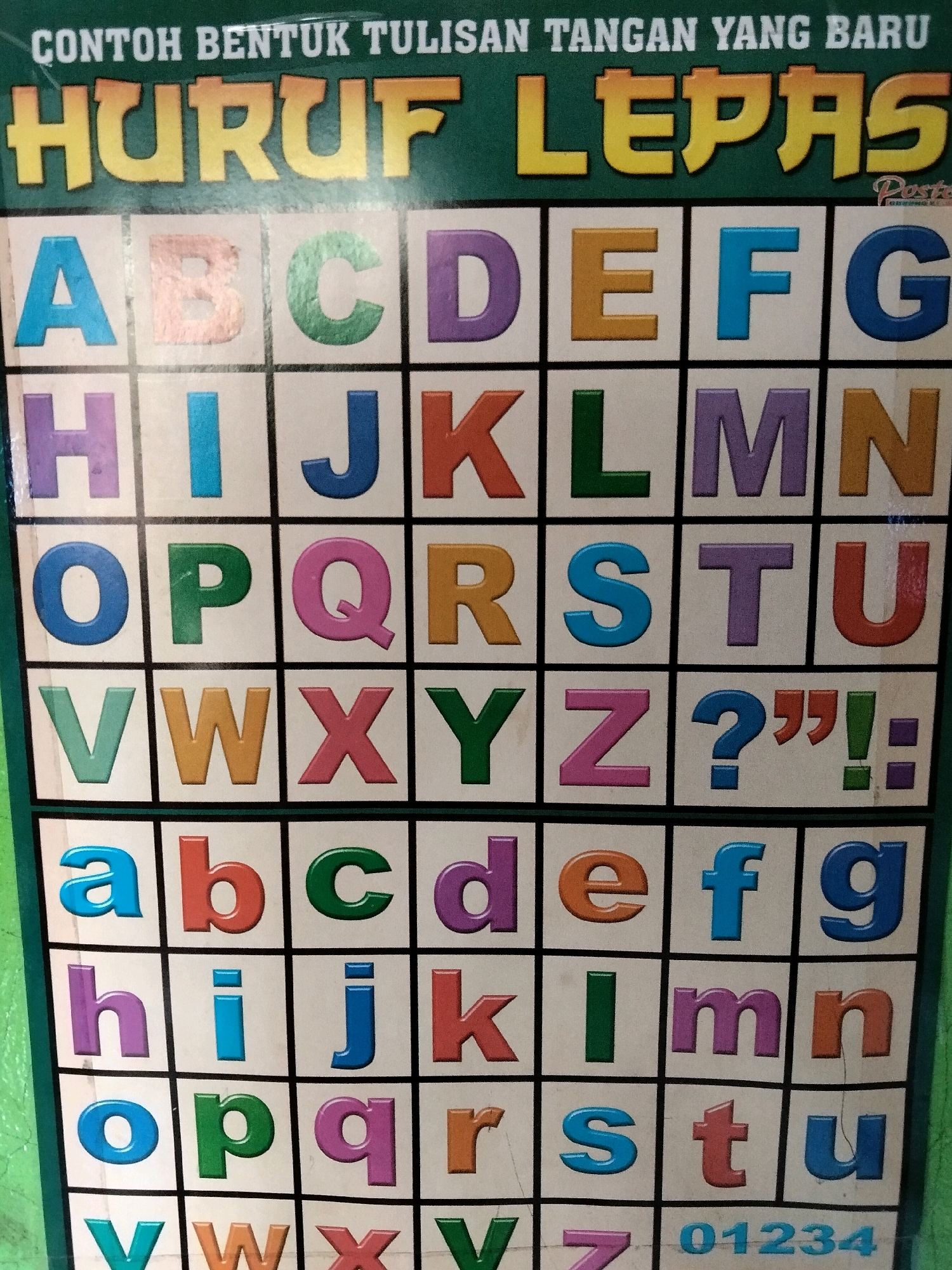 A colorful educational chart displays the Indonesian alphabet in large, bold, uppercase and lowercase letters, along with numbers. The chart is divided into a grid of squares, each containing a letter or number in a different vibrant color. The chart has a green border. The chart is mounted on a wall. The environment suggests a school or learning setting. The chart's caption reads "CONTOH BENTUK TULISAN TANGAN YANG BARU HURUF LEPAS" which translates to "EXAMPLE OF NEW HANDWRITING FORM LOOSE LETTERS". The image shows the full chart, but specific location details within the city of Rahong, Indonesia, cannot be determined from the visual information alone.