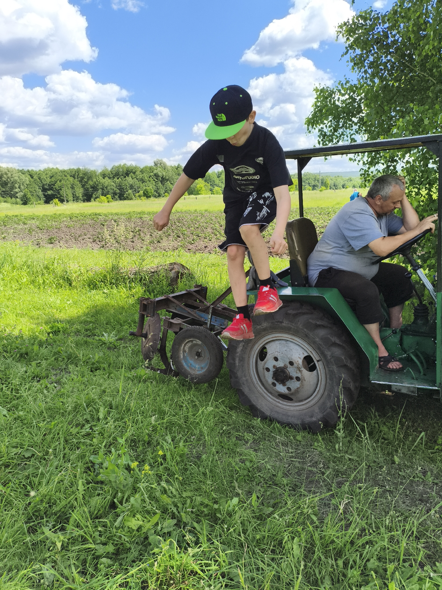 In a rural setting, likely on the outskirts of Ilmino, Russia, a boy wearing a black t-shirt, black shorts with a white pattern, a black and green baseball cap, and red sneakers stands on the back of a green tractor. He is facing away from the camera, with his right arm extended and his fist clenched. An adult male, wearing a grey t-shirt and dark shorts, sits on the tractor's driver's seat, resting his head on his left hand and holding the steering wheel with his right. He is wearing dark sandals. To the left of the tractor, a plow attachment is visible. The ground is covered in green grass, and in the background, there are fields and trees under a blue sky with white clouds.