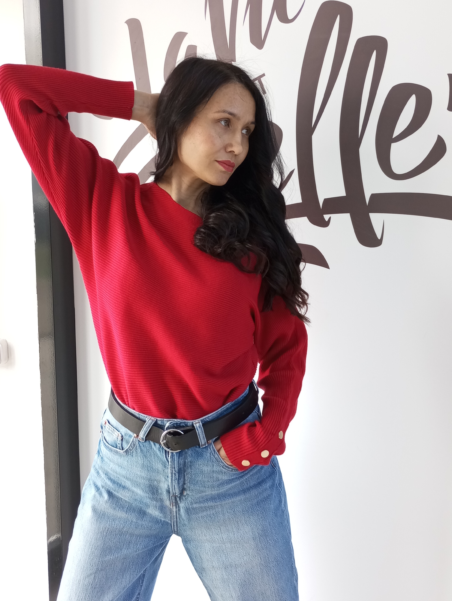 A woman with long, dark, wavy hair and visible freckles is captured in a half-body shot, posing indoors against a white wall. She is wearing a ribbed, bright red long-sleeved sweater with gold-colored buttons on the cuffs, paired with light-wash blue jeans and a black belt featuring a silver circular buckle.
Her right arm is bent, with her hand resting behind her head/neck, while her left hand is casually tucked into the pocket of her jeans. She looks towards the right side of the frame with a calm, somewhat contemplative expression. The background features large, stylized, dark brown calligraphy on the white wall, with visible letter segments that appear to form part of a word like "elle" or "Coffee." A dark vertical border or frame is present on the far left. The setting appears to be a well-lit indoor space, likely during the daytime. The location context for this image is Izhevsk, Russia.