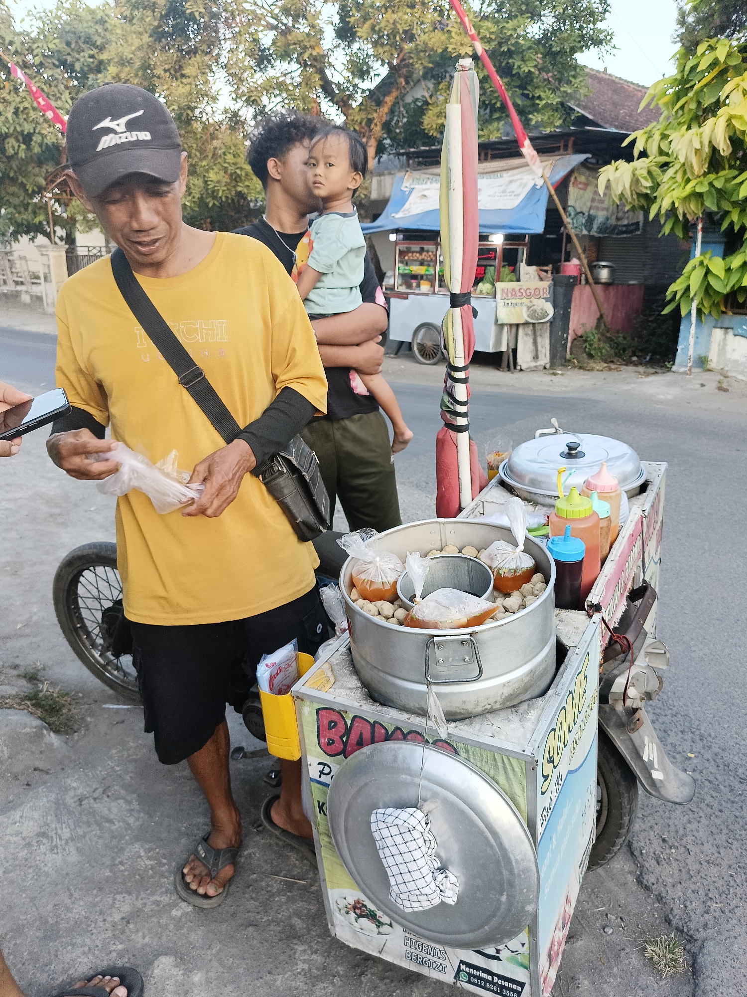 In Sukorini, Indonesia, a man wearing a yellow t-shirt, black shorts, and a black baseball cap stands by a food cart. He is holding plastic bags with what appear to be food items. Behind him, another man holds a young child. The food cart is equipped with various containers for food and condiments, including a large metal pot with a lid and several smaller bottles of sauce. The cart is mobile, attached to what looks like a motorcycle. In the background, there are buildings and trees, suggesting an outdoor setting in a residential or commercial area. A red and white pole with fabric streamers stands next to the cart. The overall scene depicts a street food vendor in action, serving customers or preparing for business.