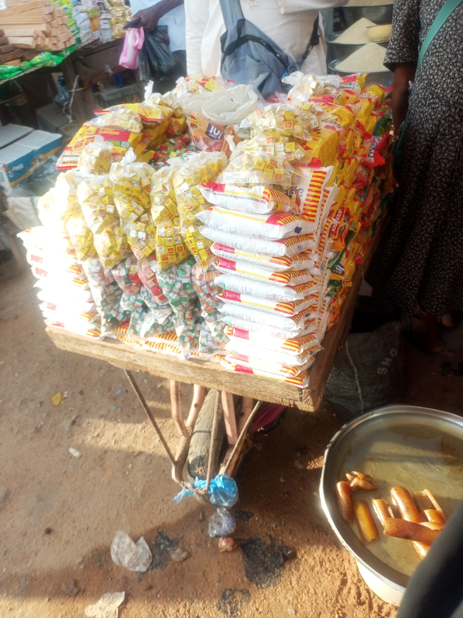 In Jalingo, Nigeria, a market stall displays a substantial quantity of packaged goods, primarily bouillon cubes and other food seasoning products, neatly stacked on a wooden cart. The products are predominantly in yellow, red, and white packaging, suggesting a vibrant and diverse selection. In the background, hints of other market items like bundles of wood and sacks of what appears to be rice are visible, along with people engaged in market activities, one holding a pink plastic bag and another in a patterned dress.
A person's leg and foot are visible in the lower right, wearing brown sandals. A large silver bowl filled with a yellowish liquid and several pieces of what look like fried meat or sausages sits on the ground to the right of the cart. The ground is sandy, indicative of an outdoor setting, and scattered debris like plastic bags and crumpled paper add to the market's atmosphere. The lighting suggests it is daytime, possibly mid-morning or afternoon, with sunlight casting distinct shadows. The overall impression is of a bustling local market scene in Nigeria, with a focus on food products and daily commerce. Some text is visible on the packaging, including "CUBES," "UNITB BATCH NO MC2/05/26 P.D.05/2026 B," and "AERICK."