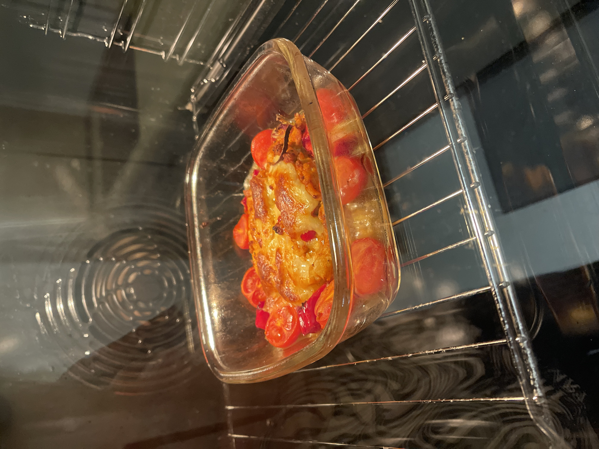 A square glass baking dish filled with food is sitting on an oven rack inside a kitchen oven. The dish contains what appears to be chicken or fish topped with melted cheese and surrounded by cherry tomatoes and possibly red bell peppers. The food is golden brown and bubbly, indicating it is hot. The oven interior is visible, including the heating element and a fan. The image is taken from a side angle, looking into the oven.