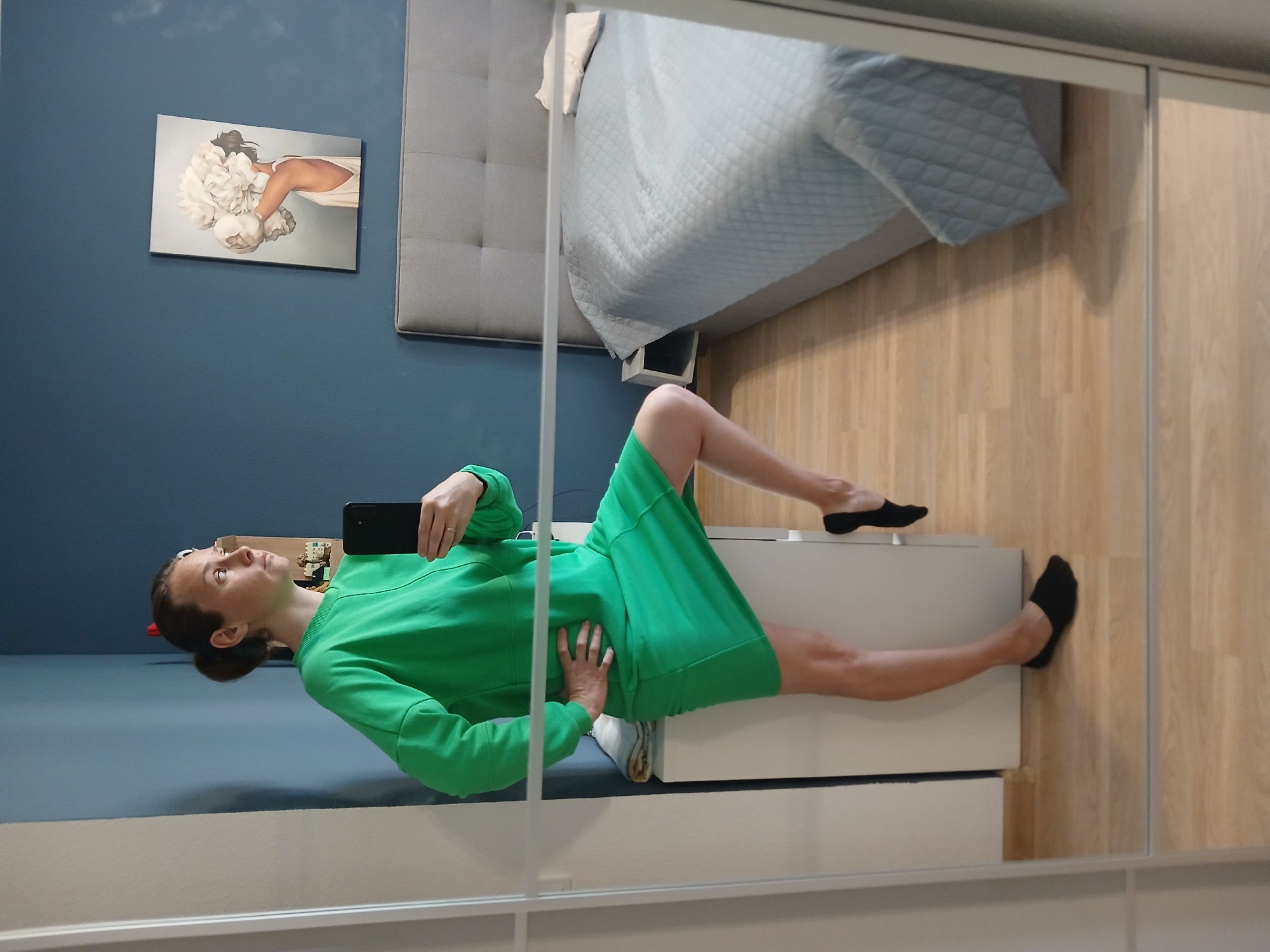 A young woman is standing in a bedroom, taking a mirror selfie. She is wearing a bright green sweatshirt dress and black socks. Her hair is pulled back in a bun. In the background, there is a bed with a gray headboard and a blue quilted bedspread. On the wall above the bed, there is a framed piece of art depicting a woman holding a bouquet of flowers. The room has light-colored wooden floors and blue walls. The image is taken from a medium shot, focusing on the woman and her reflection. The specific location in Yekaterinburg, Russia cannot be determined from the image.