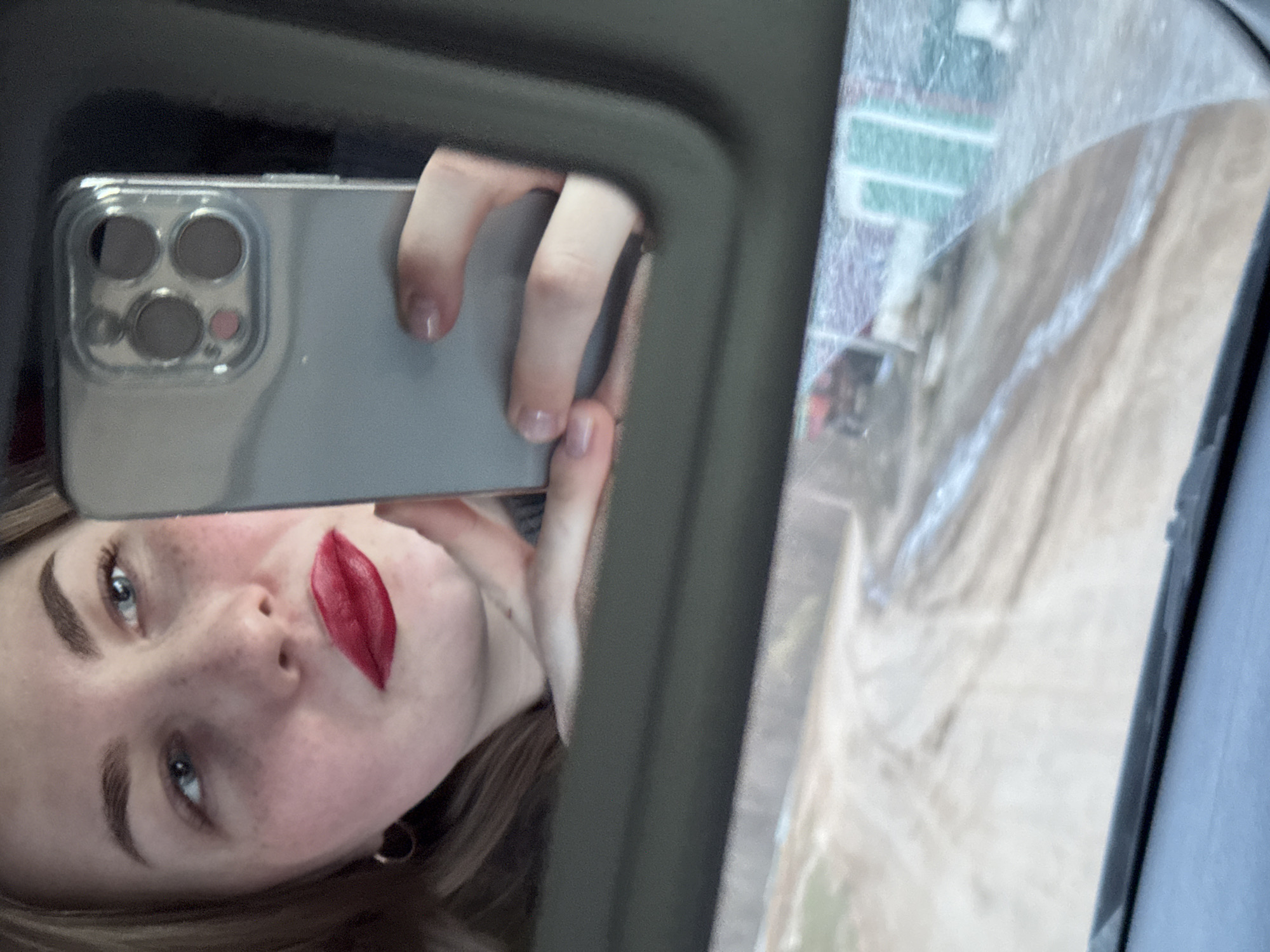 The image shows a reflection of a young woman in a mirror. The woman is holding a smartphone, presumably taking the picture. She has fair skin, freckles, and is wearing red lipstick. Her hair is light brown. The smartphone has a grey case and three camera lenses. The mirror frame is dark grey. The reflection in the mirror shows a blurry outdoor scene, possibly including some green areas and buildings. The location is Ольховка, Russia but there isn't enough information to say more about the area.