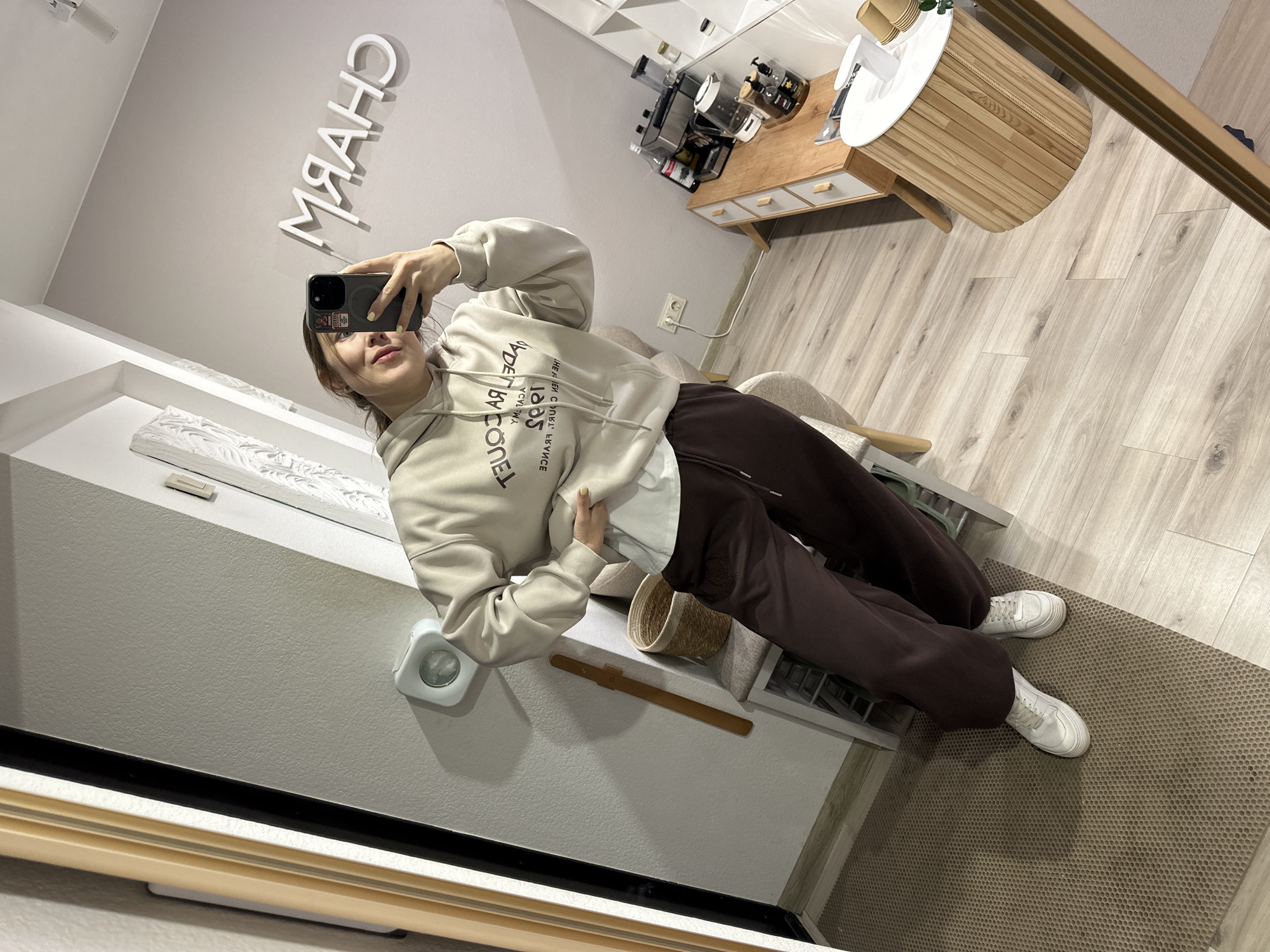 A young woman is taking a selfie in a room. She is wearing a beige hooded sweatshirt and dark brown sweatpants, with white sneakers. The room appears to be a dressing room or a bedroom. In the background, there is a mirror reflecting "CHARM" written in large white letters on the wall. There is also a wooden cabinet with drawers, a wooden side table, and various decorative items. The floor is made of light-colored wood. The woman is holding a smartphone in her right hand and posing for the photo. The overall impression is that of a casual and stylish setting.
