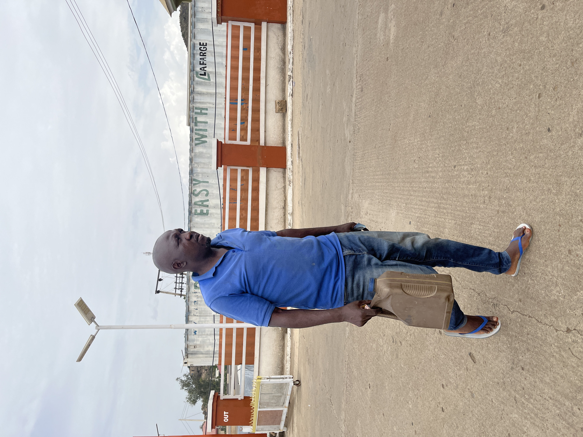 A man is standing on a paved surface. He is bald, dark-skinned, and wearing a blue polo shirt, jeans, and blue and white flip-flops. He is holding a dark plastic container. Behind him is a wall with the words "EASY WITH" and "LAFARGE" painted on it. The wall is white with orange vertical sections. In the background, there is a street lamp and power lines. The location is Gura Top, Nigeria.