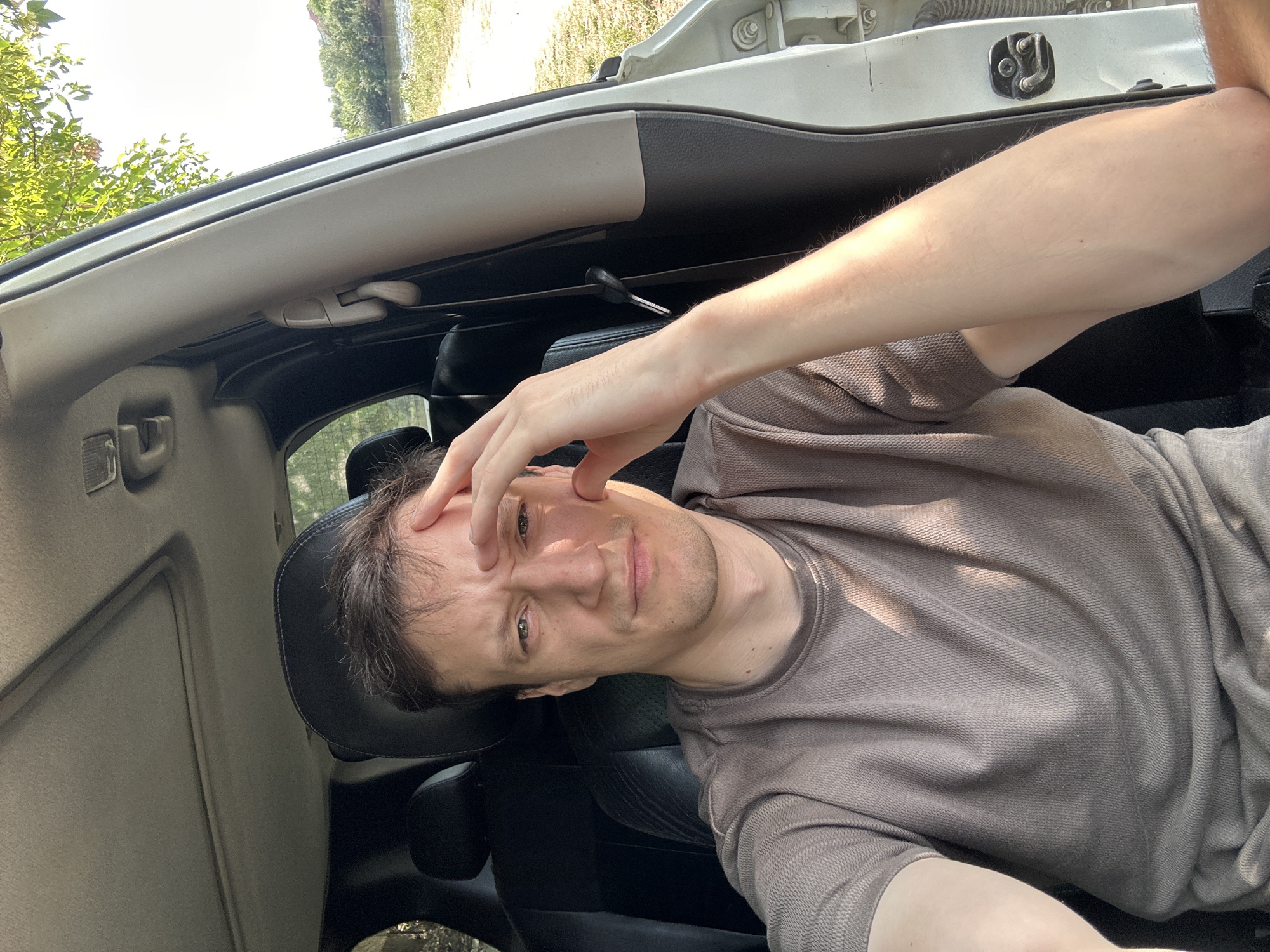 A man is sitting in the driver's seat of a car, parked outdoors. He is looking at the camera, with his right hand resting on his forehead. The car's interior is visible, including the dashboard, seats, and ceiling. Outside the car, there is greenery and a body of water, suggesting a natural setting. The light suggests it is daytime. No specific location in the city of Makiv, Ukraine can be identified from the image.