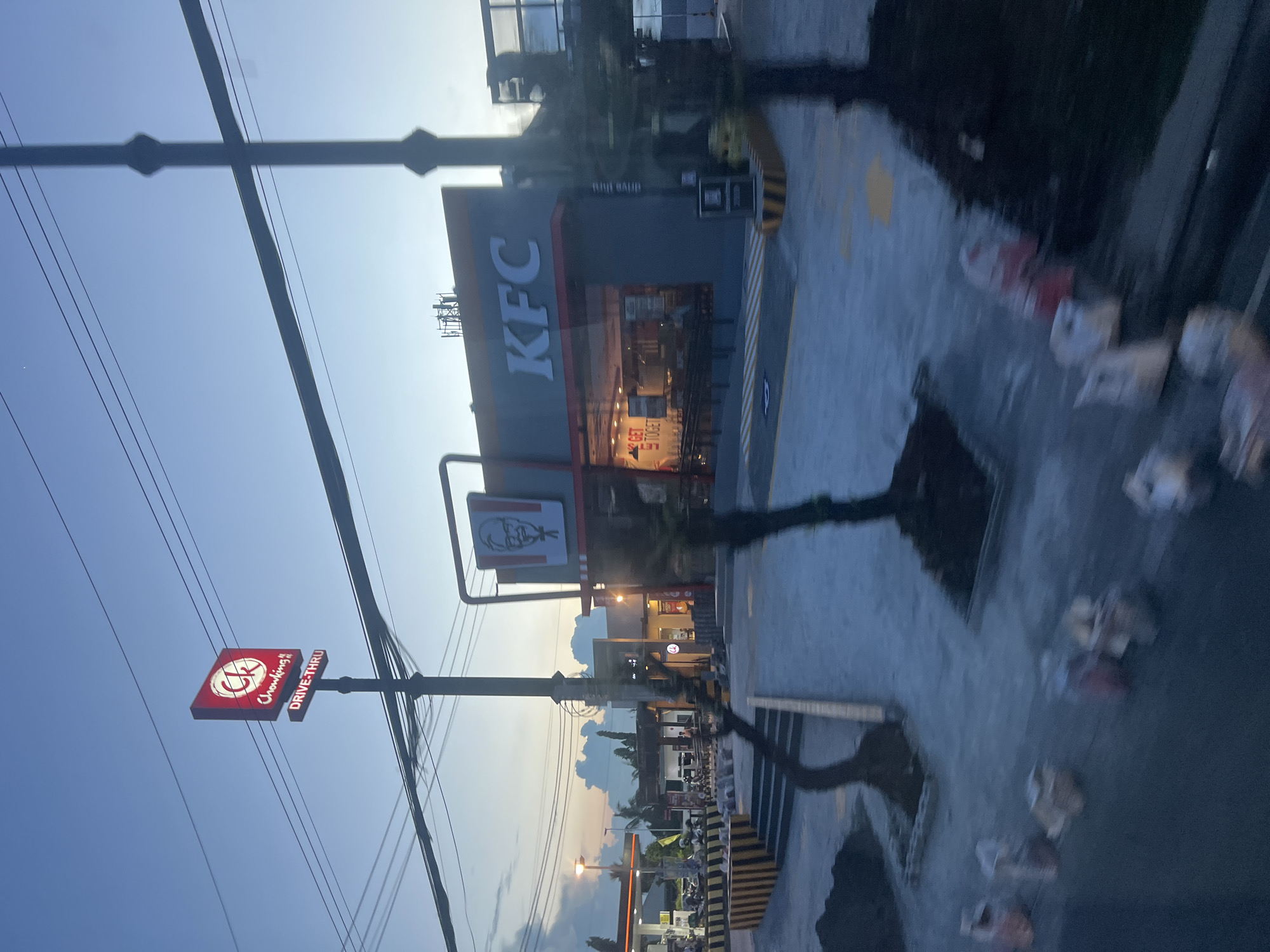 An exterior shot captures a KFC establishment in Davao City, Philippines, during twilight. The KFC building features a prominent red and gray facade with the iconic KFC logo. Adjacent to it, a Chowking sign is visible, indicating a dual-brand or nearby location. The scene is characterized by construction, with sandbags and construction materials scattered around the premises. A utility pole and power lines crisscross the sky, typical of urban Filipino landscapes. The overall atmosphere suggests a developing area or recent renovation.
