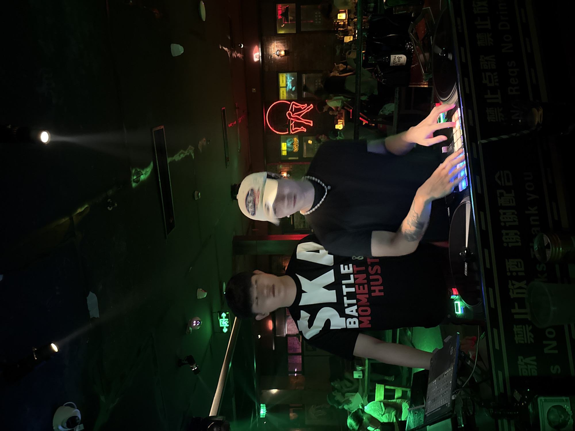 Two young Asian men are standing behind a DJ booth in a dimly lit establishment that appears to be a bar or club. The man on the left is wearing a black t-shirt with white and red lettering that reads "SKA BATTLE MOMENT & HUST". The man on the right is wearing a black t-shirt, a white baseball cap, and a necklace. He is actively operating a DJ setup, with his hands on the mixing board which is illuminated by colorful lights. In the background, other patrons are visible sitting at tables and the environment is decorated with neon lights and spotlights. The establishment appears to be located in Liangjiang, China, as indicated by the presence of Chinese text on signage and promotional materials within the venue.