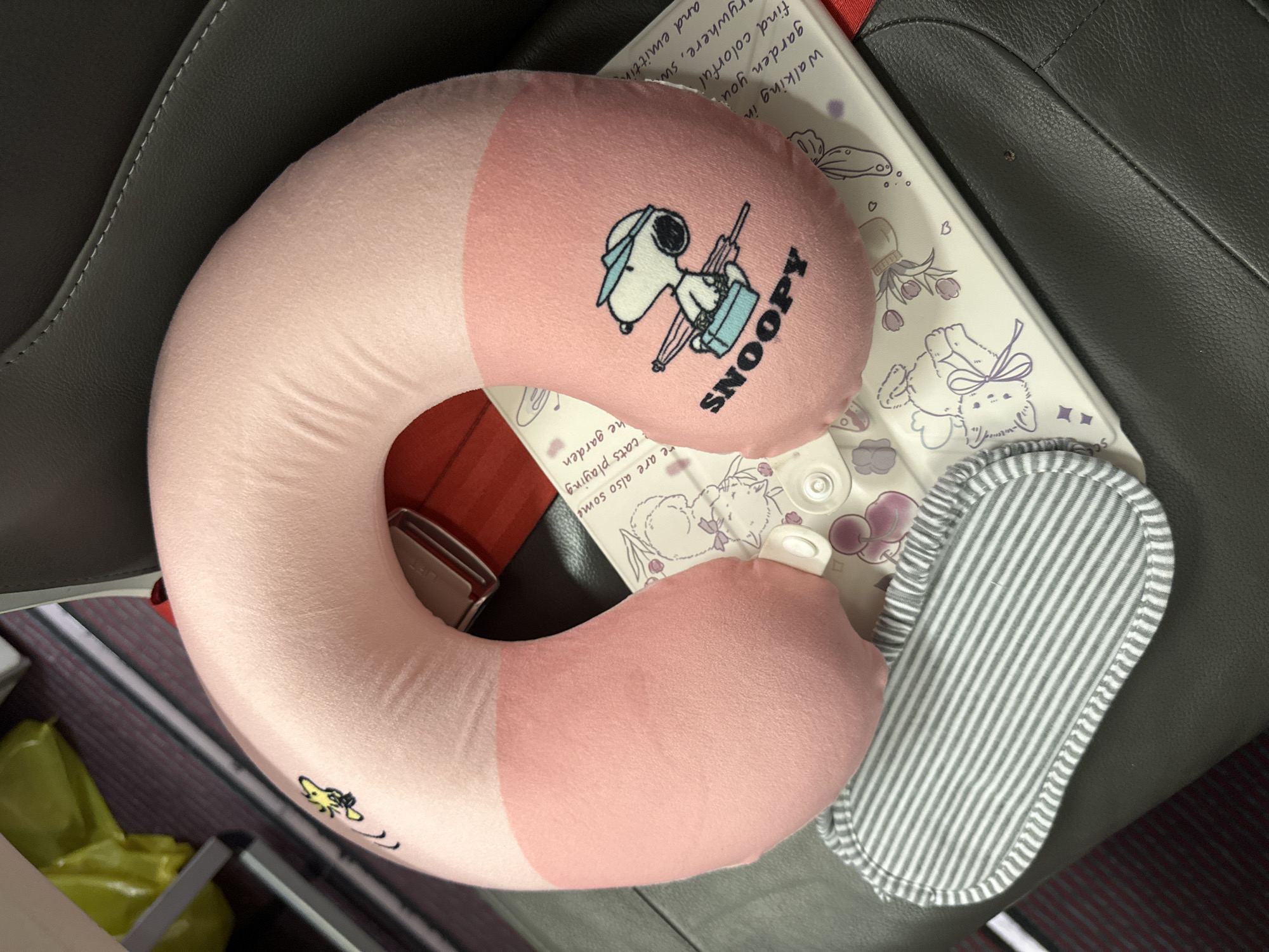 The image shows a pink and white Snoopy neck pillow on top of a white surface with drawings and text, next to a gray and white striped slipper. The scene appears to be inside a vehicle, likely a plane or bus, due to the presence of a seat belt buckle in the background. There are no people visible. The location and city of Yerevan, Armenia, cannot be determined from the provided image.