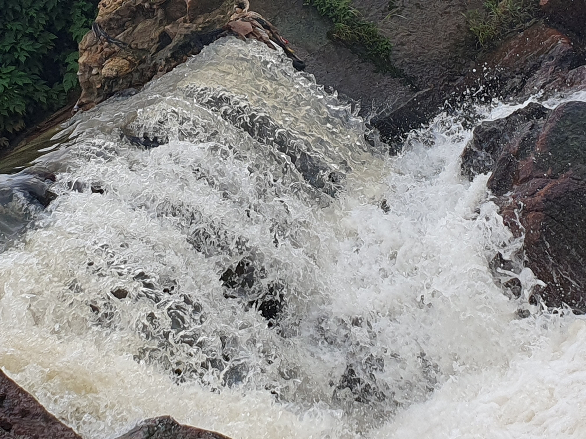 The image shows a close-up view of a waterfall cascading over rocks. The water is turbulent and foamy, with white water spraying upwards. The rocks are dark brown and reddish, with some vegetation visible on the left side of the frame. The scene is likely a natural setting, possibly in a park or wilderness area, and does not depict any urban elements within Gura Top, Nigeria. There are no people or identifiable man-made objects in the image. The focus is entirely on the movement and texture of the water and the surrounding rocks.