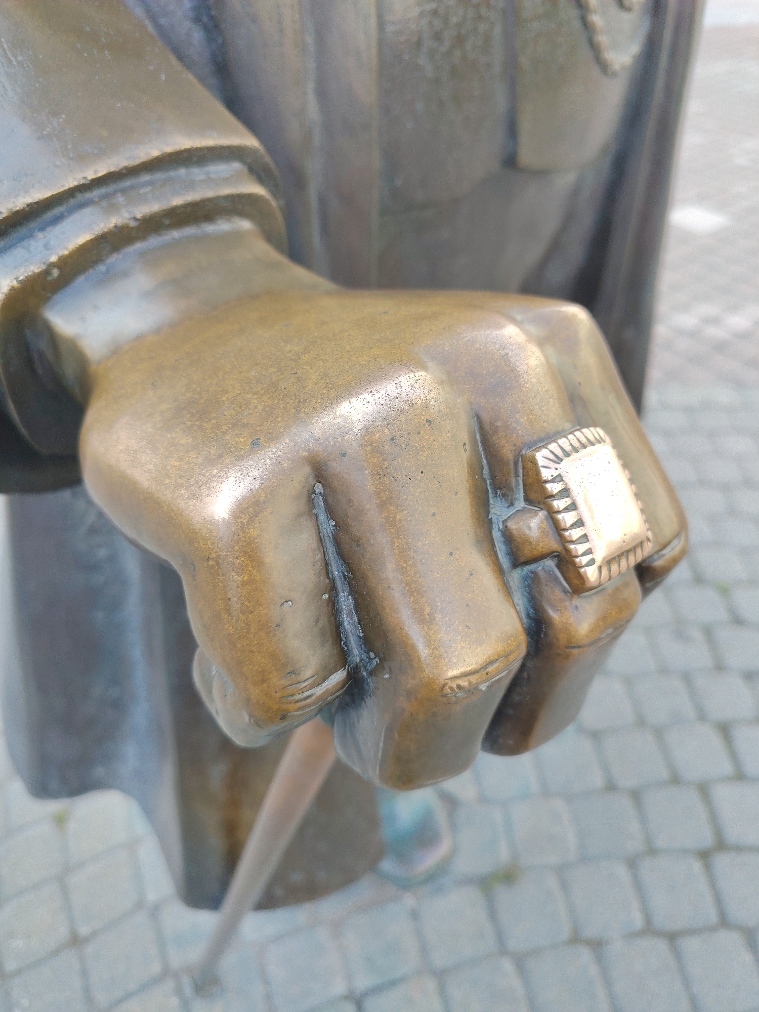 This is a close-up, eye-level, outdoor shot of a bronze statue's clenched fist. The statue is of a person, indicated by the hand and implied clothing. The fist is adorned with a ring on the index finger. The ring is square with a patterned surface and appears to be made of a lighter colored metal than the bronze of the statue. The statue's arm and body are visible behind the hand, made of bronze with a worn, aged patina. The statue appears to be holding something thin and cylindrical with its left hand, though this object is largely out of focus. The background consists of a paved surface, likely cobblestones, suggesting an outdoor public space. The lighting suggests it is daytime, with some soft shadows indicating overcast or diffused sunlight. The scene is static, depicting a detail of a larger sculpture, and evokes a sense of stillness and permanence. No text is visible in the image. The context provided indicates the location is Yekaterinburg, Russia.