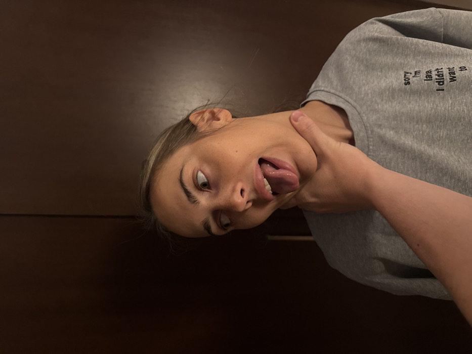 The scene is an indoor setting in Warsaw, Poland. A female individual, appearing to be an adult, is depicted from the upper chest upward, positioned against a dark brown background, likely a closet or wardrobe door. She has light skin and light-colored hair, which is pulled back. The individual wears a grey long-sleeved top featuring partially visible black text that reads 'sorry I'm late. I didn't want to'. Her face displays a wide-eyed expression, with her gaze directed towards the upper right. Her mouth is open, and her tongue is extended, suggesting a non-distressed or playful interaction. A human hand, with a lighter skin tone, is grasping her neck; the thumb is positioned on the left side, and fingers wrap around the right. The grip appears to be light. The overall lighting is dim, emanating from the front.