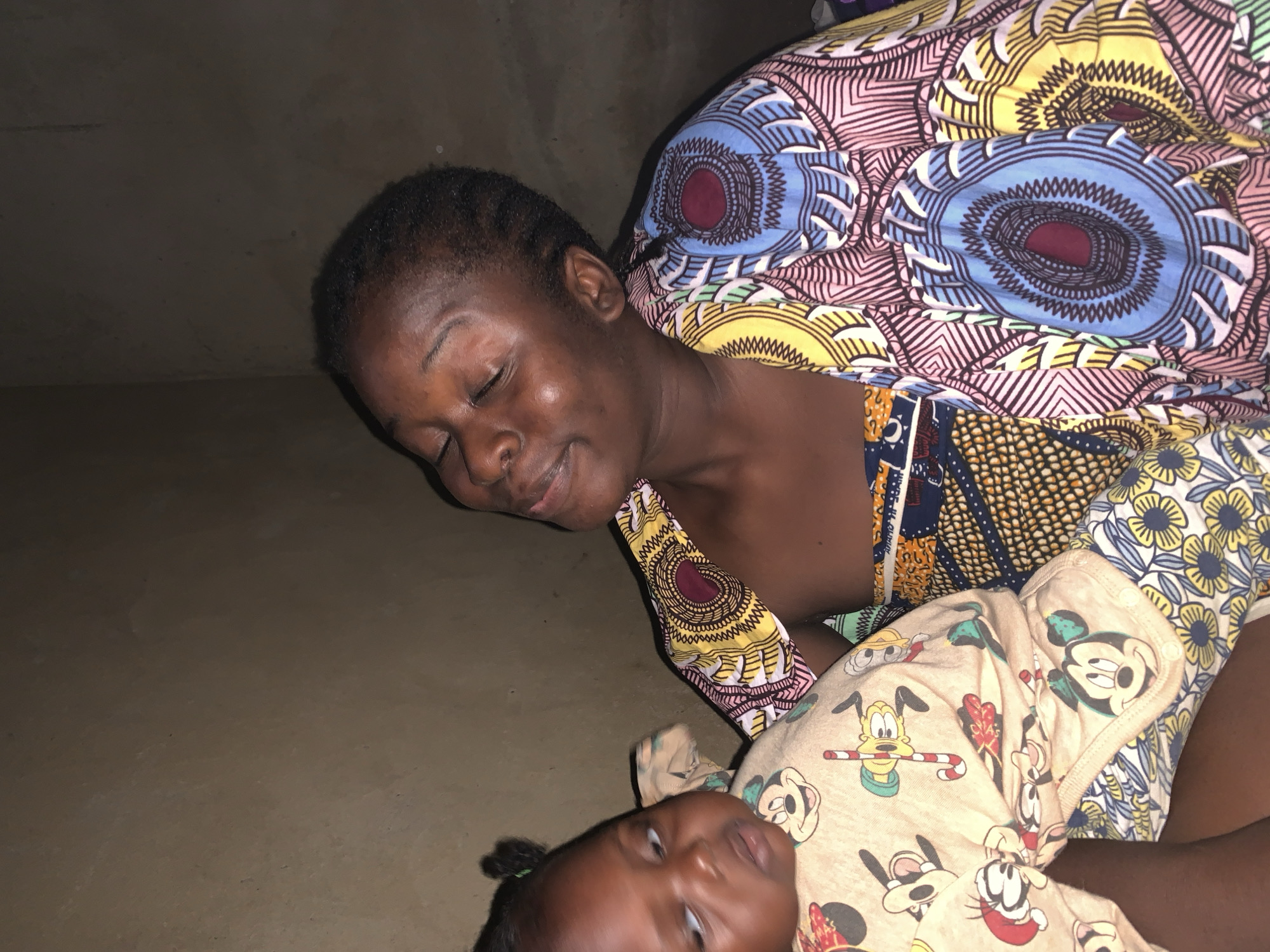 In the city of Agban, Nigeria, a young woman cradles a baby. The woman, with her eyes closed, appears to be smiling. She is wearing a vibrantly patterned cloth. The baby, whose eyes are open and looking to the side, is dressed in a light-colored garment adorned with cartoon characters, including Mickey Mouse. The background is a textured, dark brown wall.