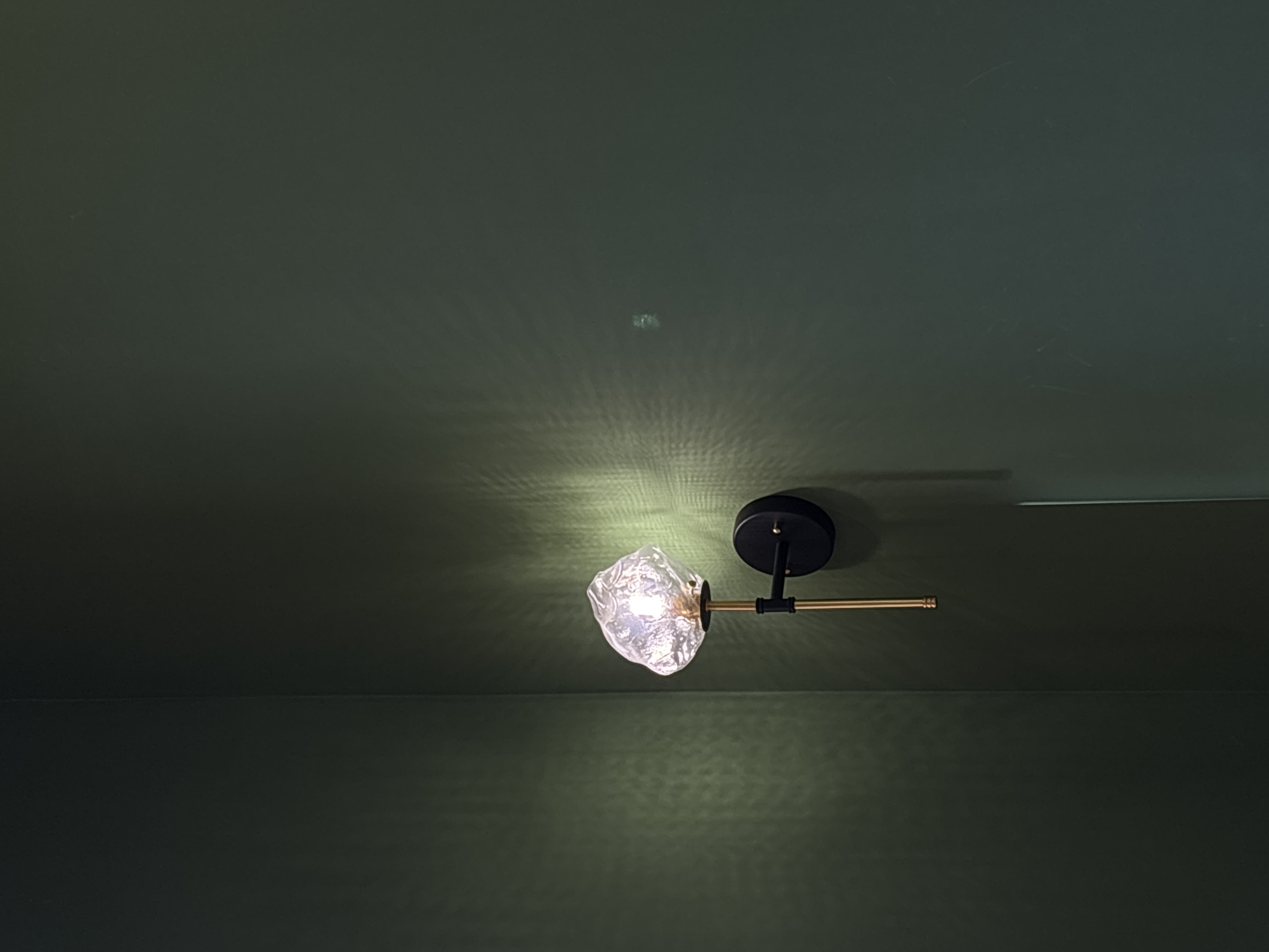 The image shows a modern wall-mounted light fixture against a dark green wall. The fixture has a black circular base, a gold-colored arm extending from the base, and a textured, possibly glass, lampshade that emits a bright light. There is a horizontal line suggesting a wall division. The exact location within Chaikovsky, Russia, is not discernible from the image. No people, subjects, or specific actions are visible.