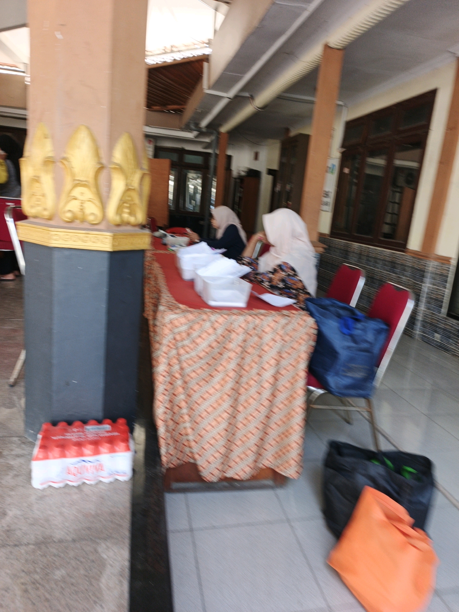 The image depicts an outdoor scene in Ketegan, Indonesia, possibly a communal area or event space. In the foreground, a tall, square pillar with ornate gold decorations is visible on the left. To the right of the pillar is a table covered with a patterned orange and white cloth. On the table are several white plastic containers.
Two individuals are present. One, wearing a light-colored hijab, is seated at the table, appearing to be engaged in an activity. Another person, also wearing a hijab, is partially visible in the background, further to the left.
In front of the table, to the left, is a package of bottled water, with red and white packaging. To the right of the table, two red chairs are positioned. On one chair is a dark blue bag. On the floor in front of the chairs are two more bags: one black and one orange. The ground surface appears to be tiled. The background shows a building with dark-framed windows and an overhang. The overall impression is a functional, possibly temporary, setup for an event or gathering.