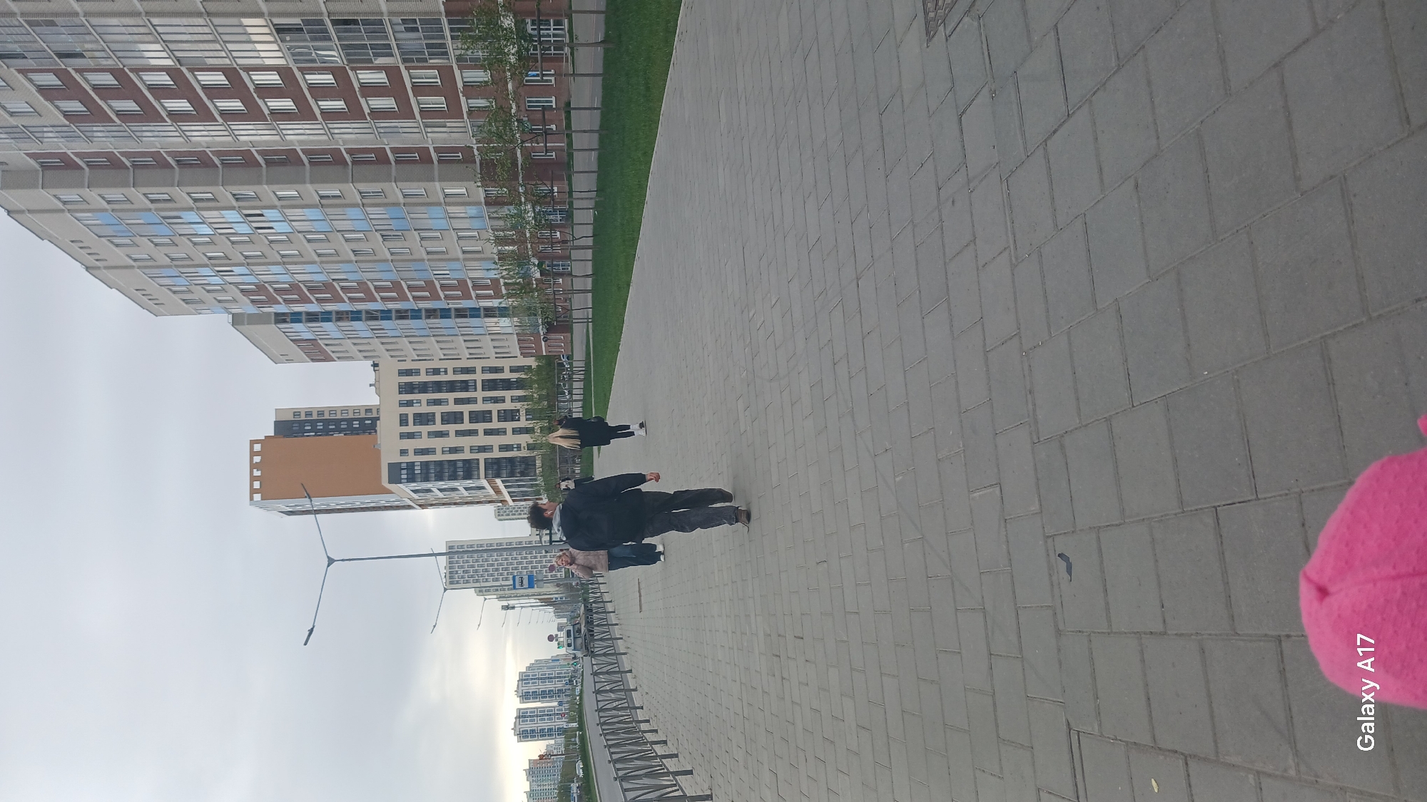 The image captures a street scene in Yekaterinburg, Russia. A wide sidewalk composed of gray paving stones stretches into the distance. A man in a dark jacket and gray pants is walking along the sidewalk, accompanied by a woman and child. Further ahead, another woman with blonde hair is also walking. Tall apartment buildings dominate the left side of the frame. The buildings are of varying heights and architectural styles, including a structure with red brick accents. Street lights are visible along the road. The sky is overcast. A corner of pink material is visible in the lower right, obscuring part of "Galaxy A17" text.