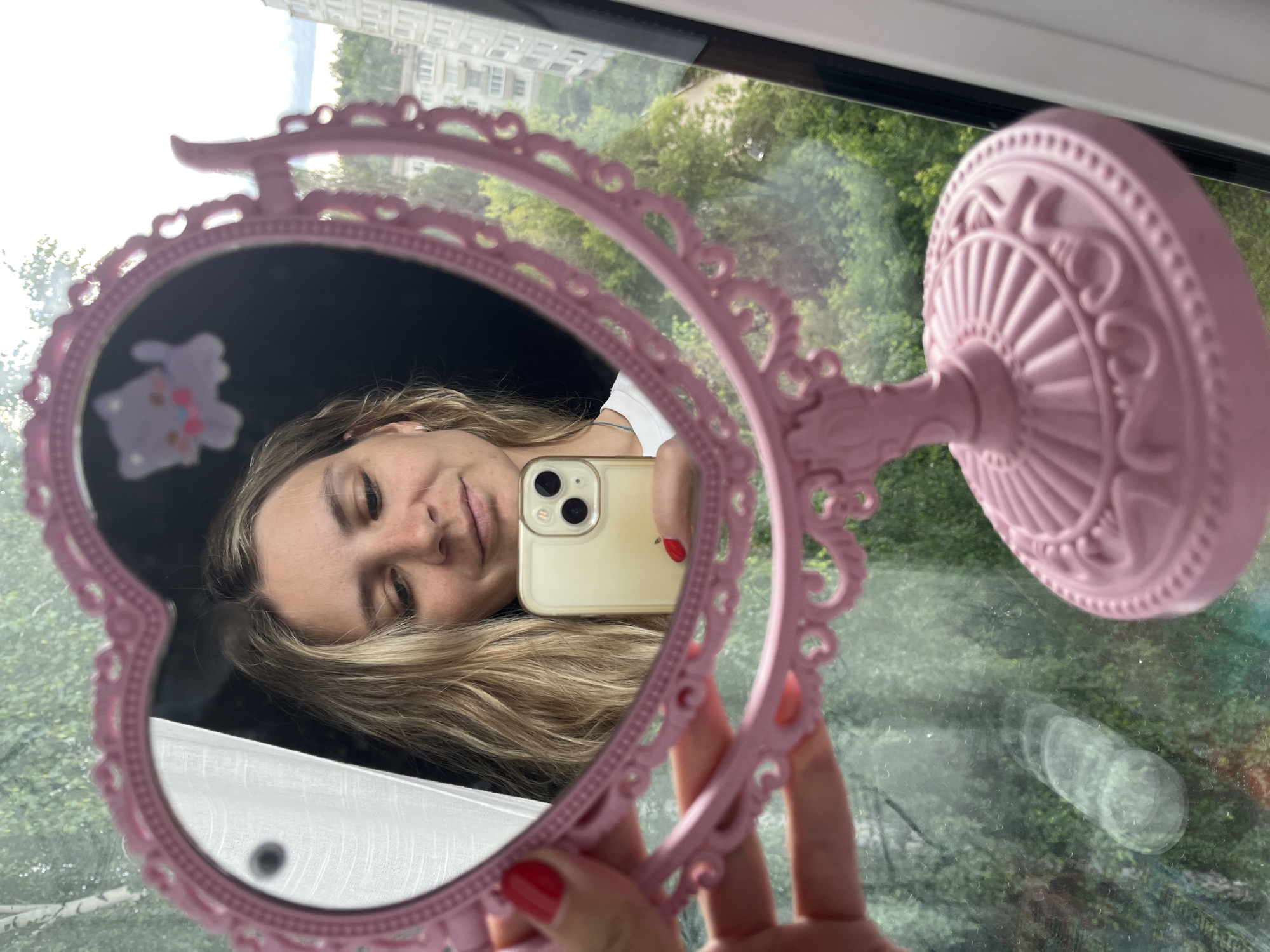 A woman takes a selfie in a pink heart-shaped mirror. Her face is reflected in the mirror, with her blonde hair falling over her shoulder. She is holding a white iPhone in her right hand, and her nails are painted red. In the reflection, a small purple bunny sticker is visible on the black backing of the mirror. The background of the mirror's reflection shows greenery, suggesting an outdoor scene. The mirror itself has decorative carvings and sits on a stand, also pink. The overall setting appears to be indoors, near a window, with sunlight illuminating the scene. The woman's expression is relaxed and pleasant. The location is identified as Chaikovsky, Russia, implying the window looks out onto a view typical of that region.