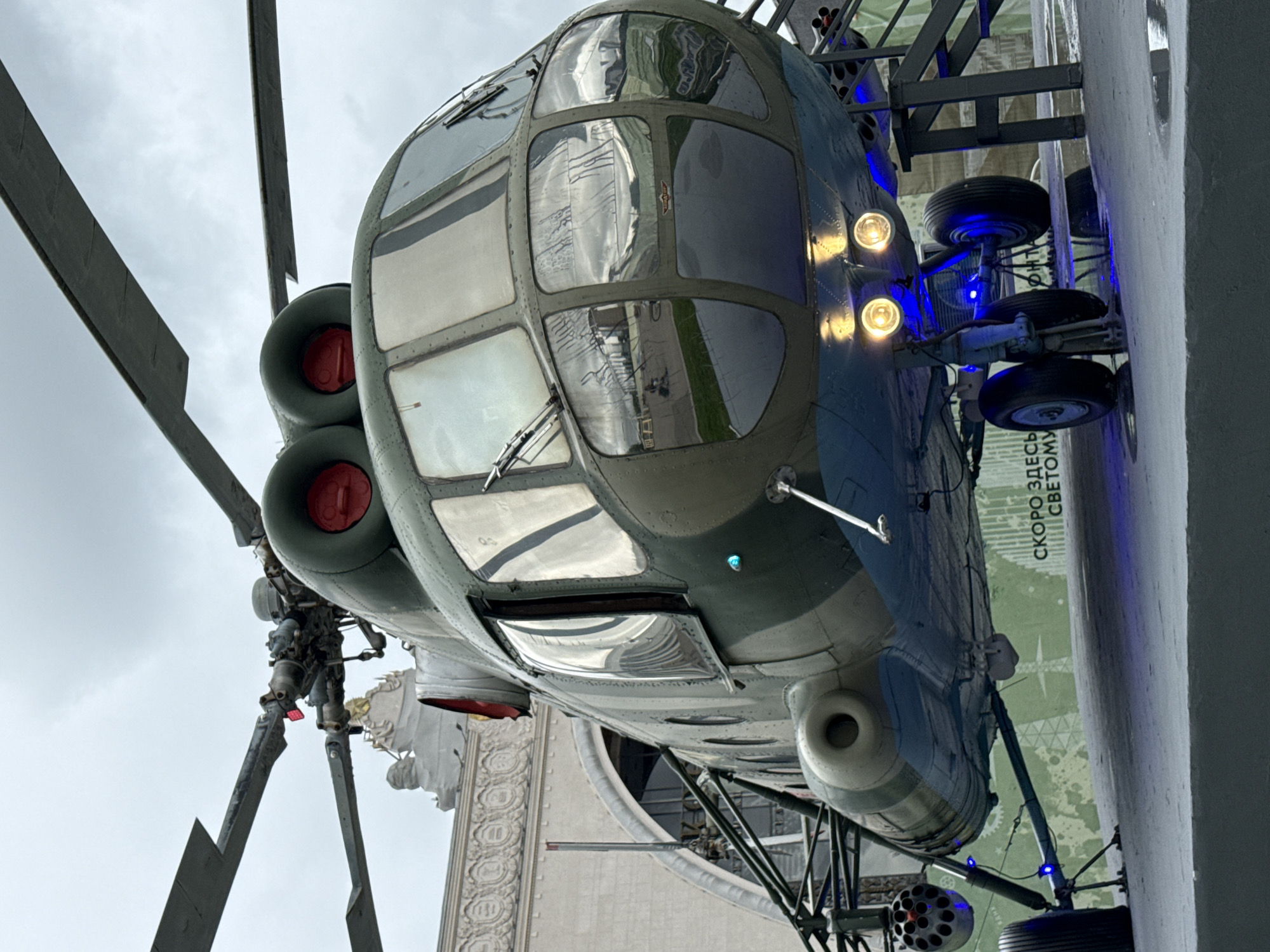 This is a low-angle, full-shot, eye-level photograph of a parked Soviet-era Mil Mi-8 helicopter, a military transport model. The helicopter is painted in a camouflage scheme of dark green and grey, with a blue underside. It is positioned on a raised platform, with its landing gear visible. The front of the helicopter features a cockpit with multiple windows that reflect the cloudy sky and surrounding environment. Two large engines are visible at the top of the helicopter, each with a red circular intake. The helicopter has rotor blades above, with the tips extending out of the frame.
To the left of the helicopter, a rotary cannon pod is attached to the fuselage. The helicopter is illuminated by bright yellow lights mounted underneath the fuselage and blue accent lights at the front landing gear and on the side of the aircraft. In the background, a large, ornate, pale-colored building with arched windows and decorative architectural elements is visible. The building appears to be made of concrete or stone. On a wall behind the helicopter, there is text in Russian, which appears to read "СКОРО ЗДЕСЬ СВЕТОМУ" (Soon here is light). This text suggests the location might be an exhibition or museum related to aviation or technology.
The photograph was taken at the Moscow Exhibition of Achievements of National Economy (VDNKh), a large public exhibition and amusement park in Moscow, Russia. The helicopter is part of the "Cosmos" pavilion, which showcases the history of Soviet and Russian space exploration and aviation. The weather appears to be overcast. There are no people visible in the photograph.