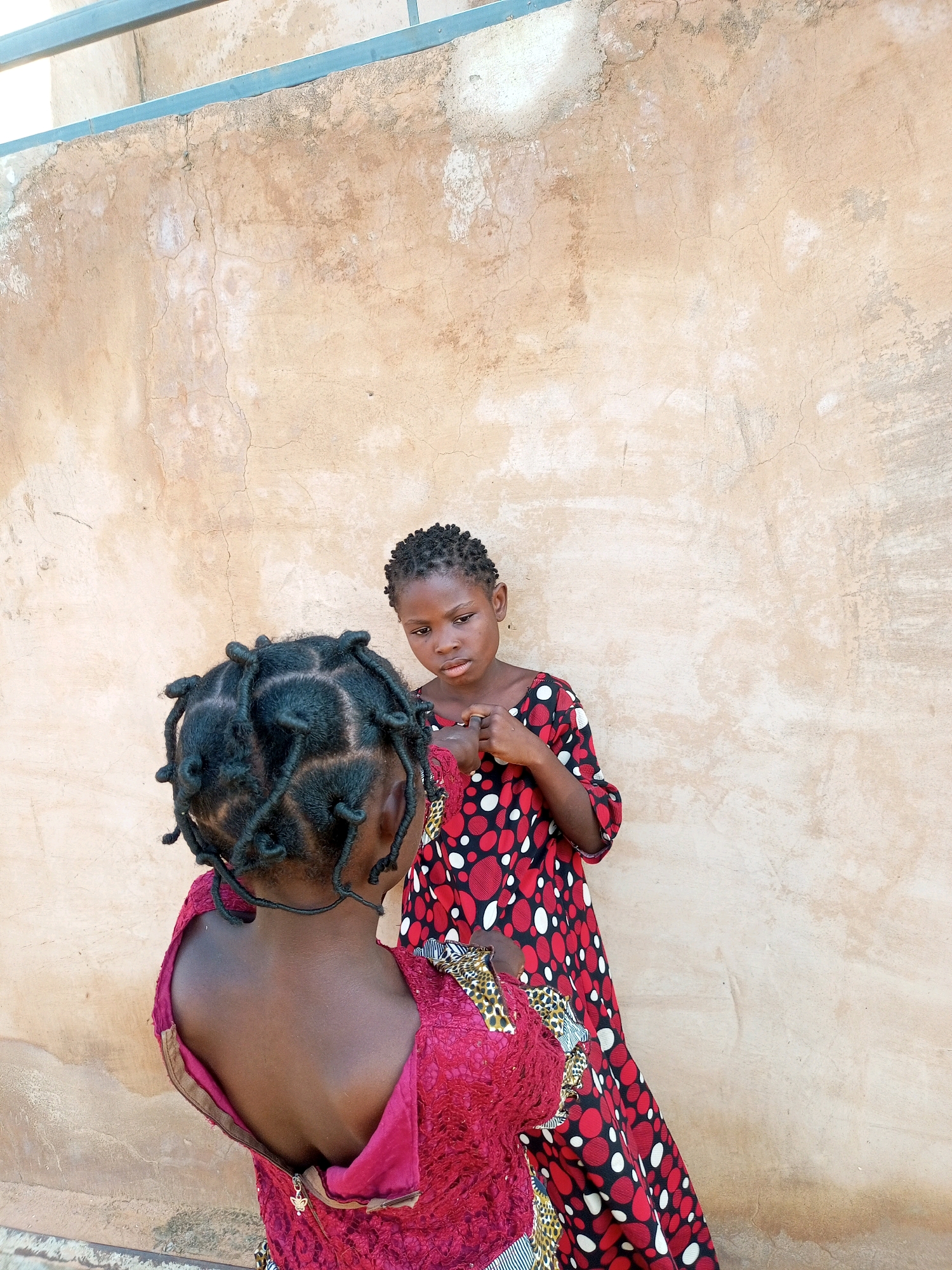 In a dusty, outdoor environment in Tumfure, Nigeria, two young girls stand against a textured, light-brown stucco wall. The girl in the foreground, facing away from the camera, has her dark hair styled in numerous small, rounded braids. She is wearing a dark pink, possibly lace, dress with an open zipper in the back. To her right, the second girl, facing the camera, has her dark hair also styled in short braids. She is wearing a vibrant red dress adorned with a pattern of white and black polka dots. Her hands are clasped together in front of her. The wall behind them appears weathered and uneven, with visible cracks and variations in color. A narrow metal bar runs horizontally across the top of the wall.
