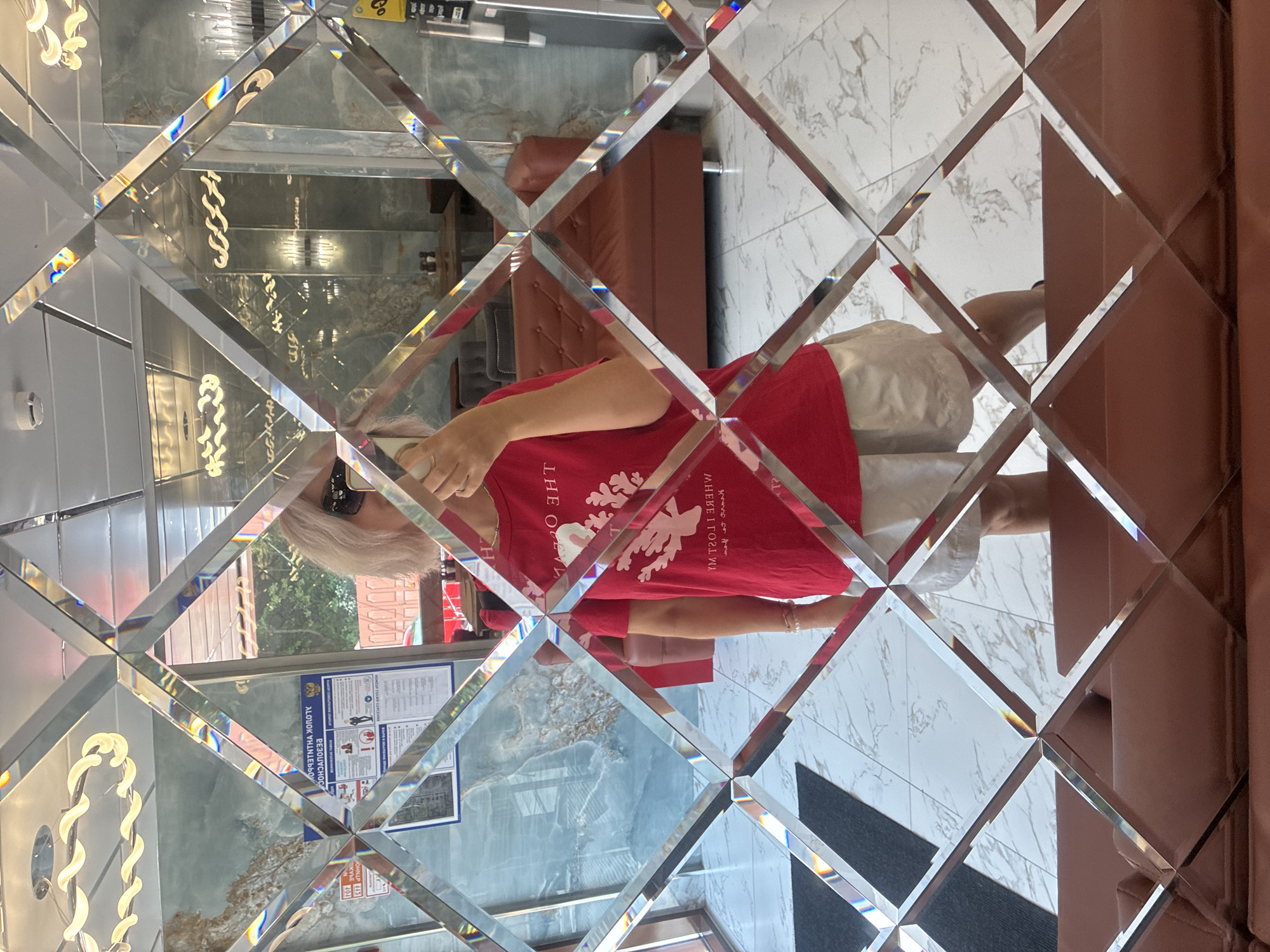 The image shows a person taking a selfie in a mirrored room. The person is wearing a red t-shirt with a white design and light-colored shorts. The room has a tiled floor, a marbled wall, and several couches. A poster with Russian text is visible on the wall. The ceiling has decorative lighting fixtures. The mirror is made of many small, diamond-shaped panes, creating a fragmented reflection of the scene. The location is stated as Estosadok, Russia.