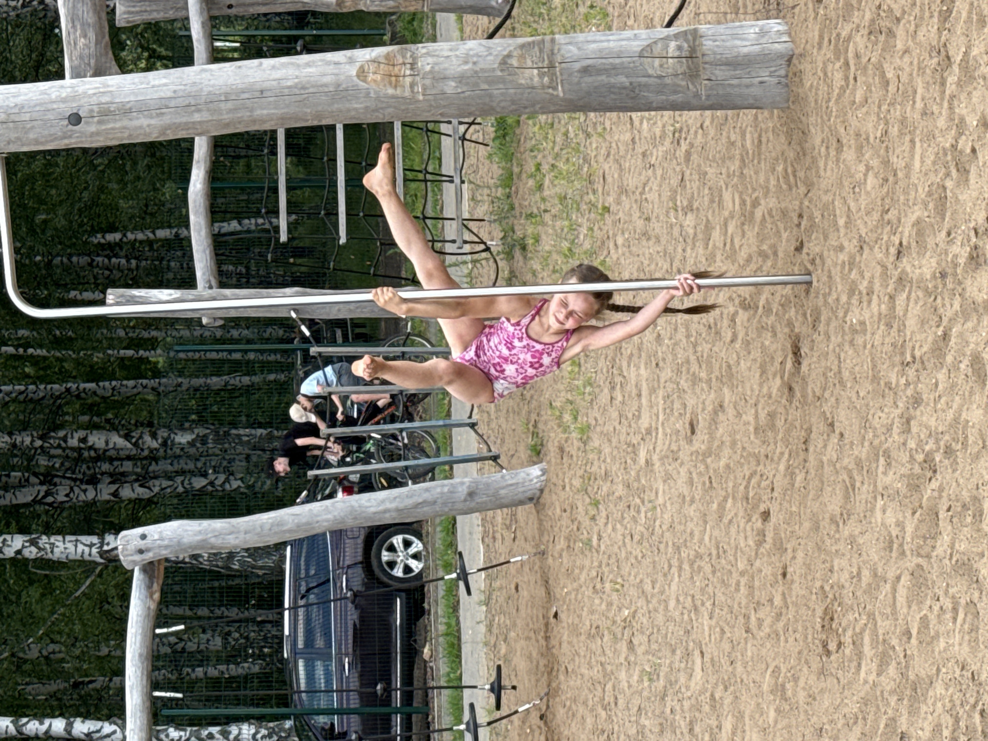 The image shows a young girl in a pink swimsuit performing a gymnastic move on a horizontal bar at an outdoor playground. She is upside down, with her legs extended and her arms gripping the bar. Her body is curved, and she appears to be in mid-air. She has a determined expression on her face, with her eyes closed and a slight smile. Her braided brown hair is flying out behind her.
The playground equipment is made of natural materials, with thick wooden logs forming the structure. The ground beneath the bar is covered in sand. In the background, there is a dense forest with birch trees, and a dark-colored car parked on a paved surface. Several other people are visible in the background, some appearing to be watching the girl. There is also a rack of bicycles. The lighting suggests it is daytime, with soft, diffused light, possibly indicating an overcast or cloudy day. The overall scene conveys a sense of athleticism and playfulness in a natural setting.