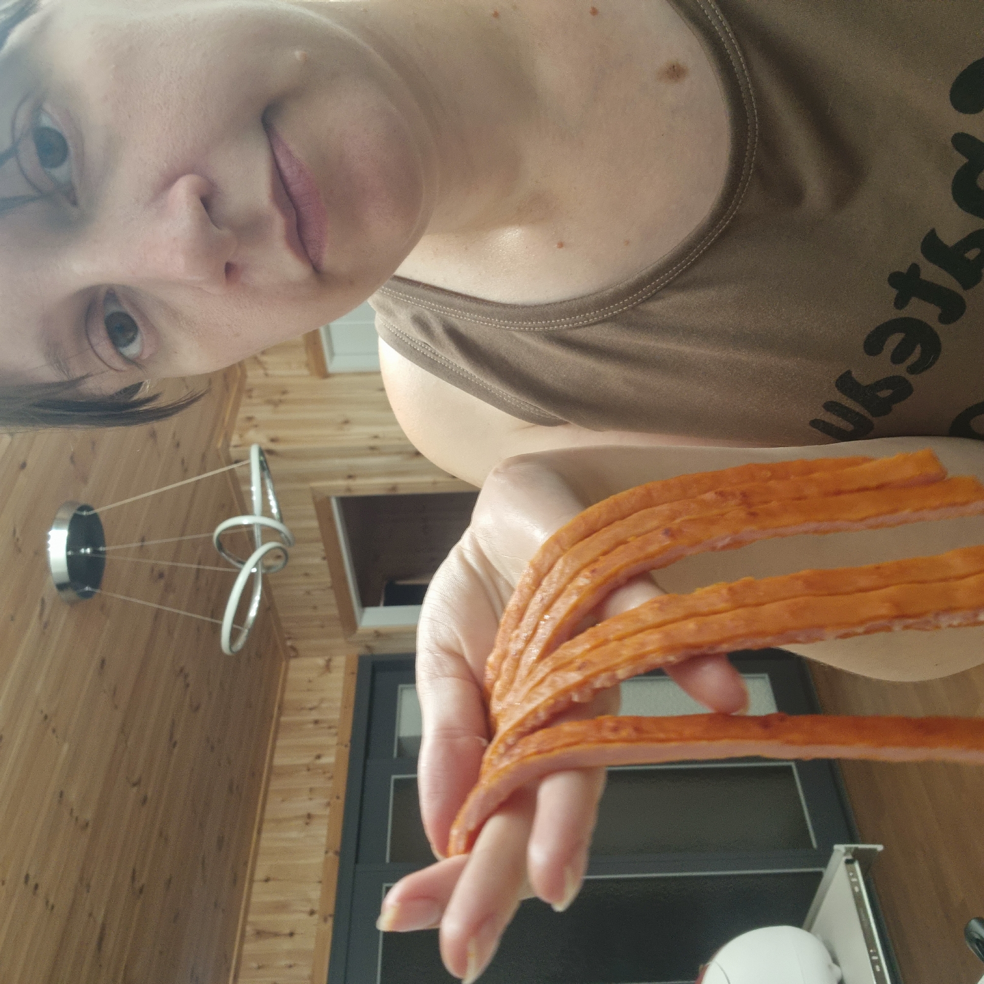This is a selfie, likely taken indoors given the wooden wall paneling and ceiling light fixture visible in the background. The image is rotated so that the subject's face, which is partially visible at the top of the frame, appears upside down. The subject is holding a fan of sliced meat, possibly hot dogs or a similar processed meat product, in their hand, which is positioned in the lower right quadrant of the image. The meat slices are thin and have an orange-pink hue. The subject is wearing a brown tank top with visible stitching along the neckline and shoulder straps. The skin of the subject shows a few small moles. The lighting suggests it could be daytime, as there is some natural light visible. The overall scene appears casual and domestic. There is text on the subject's tank top, appearing to be a stylized font, but it is partially obscured and difficult to read fully.