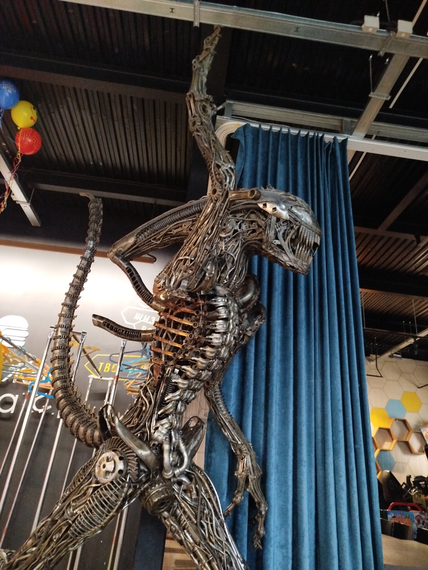 This image shows a metallic sculpture of an alien creature, resembling the Xenomorph from the "Alien" franchise, prominently displayed indoors. The sculpture is intricate and appears to be constructed from various metal parts, showcasing a detailed, skeletal, and mechanical aesthetic. It is positioned in front of a dark blue, pleated curtain, and partially obscured by a black ceiling structure with exposed metal beams and wiring. In the upper left corner, three balloons are visible: one blue, one yellow with a smiley face drawn on it, and one red with white polka dots. In the background, to the right of the curtain, a wall with hexagonal tiles in yellow, blue, and white is visible, along with some shelving and what appears to be a play area or arcade with colorful items and a monitor. To the left of the sculpture, some brightly colored metal structures and a dark wall with white text are partially visible. The overall impression is of a decorative display, possibly in a themed establishment or event space. There is no indication of the city of Mirny, Russia, in the visual elements of the image.