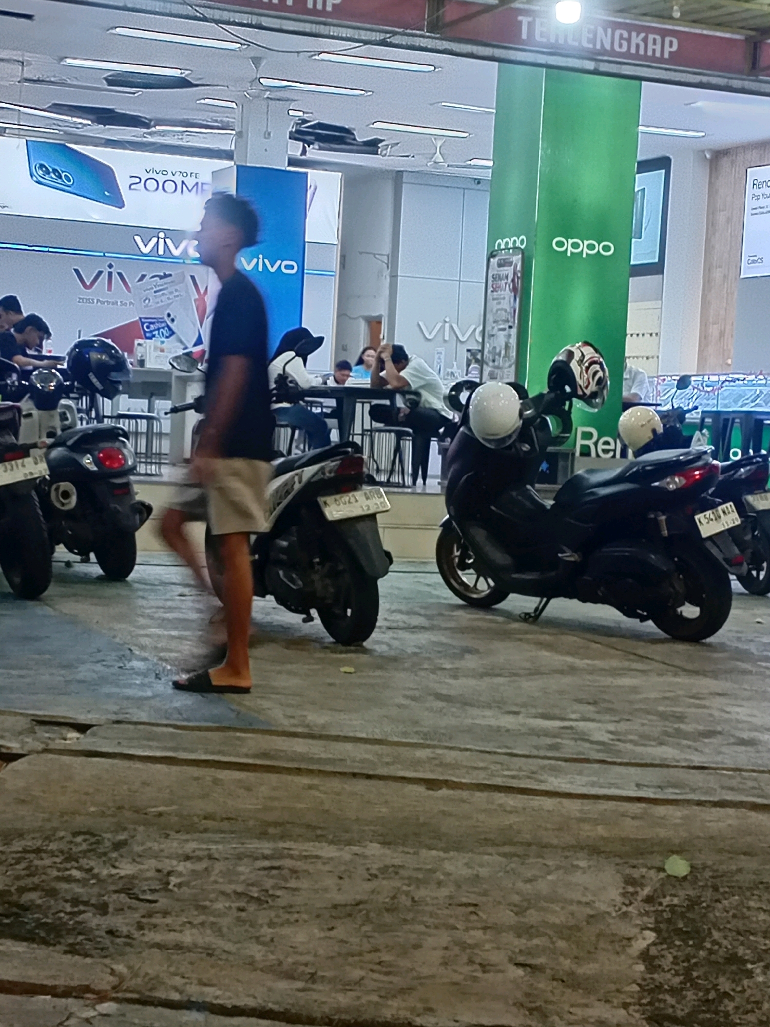 The image depicts a street scene in Kudus, Indonesia, likely in front of an electronics store. In the foreground, several parked motorcycles are visible, some with helmets placed on them. A person, seen from the waist down, is walking, wearing dark shorts and sandals. In the background, there are large advertisements for mobile phone brands like "vivo" and "oppo." People can be seen sitting at tables behind the motorcycles, suggesting an outdoor seating area or a waiting area associated with the store. The environment appears to be an urban setting with commercial signage and a paved ground surface. The lighting suggests it is daytime.