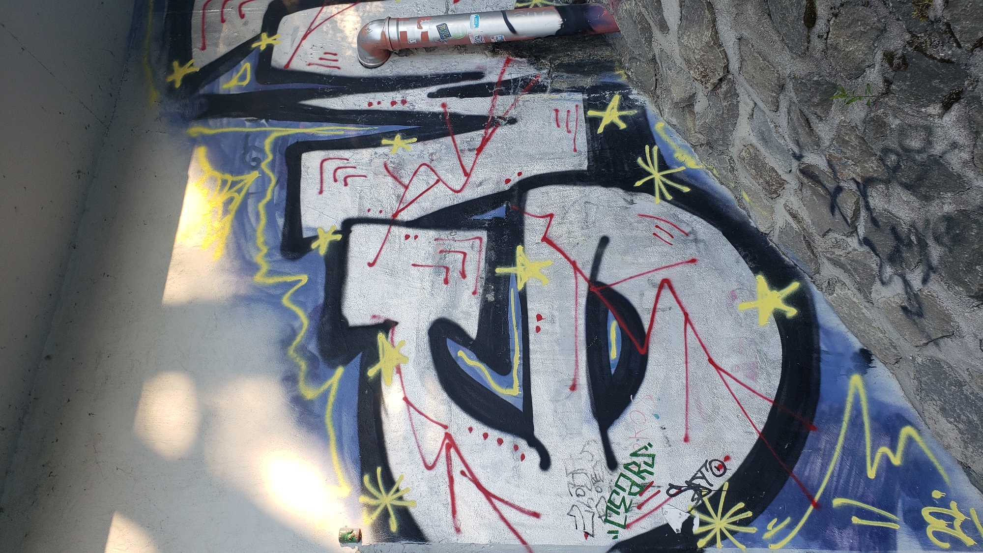 The image depicts a section of a concrete wall with graffiti in Prešov, Slovakia, near a stone embankment. The graffiti is rendered in silver, black, blue, and yellow, with red lines and small yellow star shapes scattered across the wall. A silver pipe with some stickers is visible on the right side of the graffiti. The wall is illuminated by sunlight casting shadows. The stone embankment is visible at the bottom of the image, with a small plant growing from it.