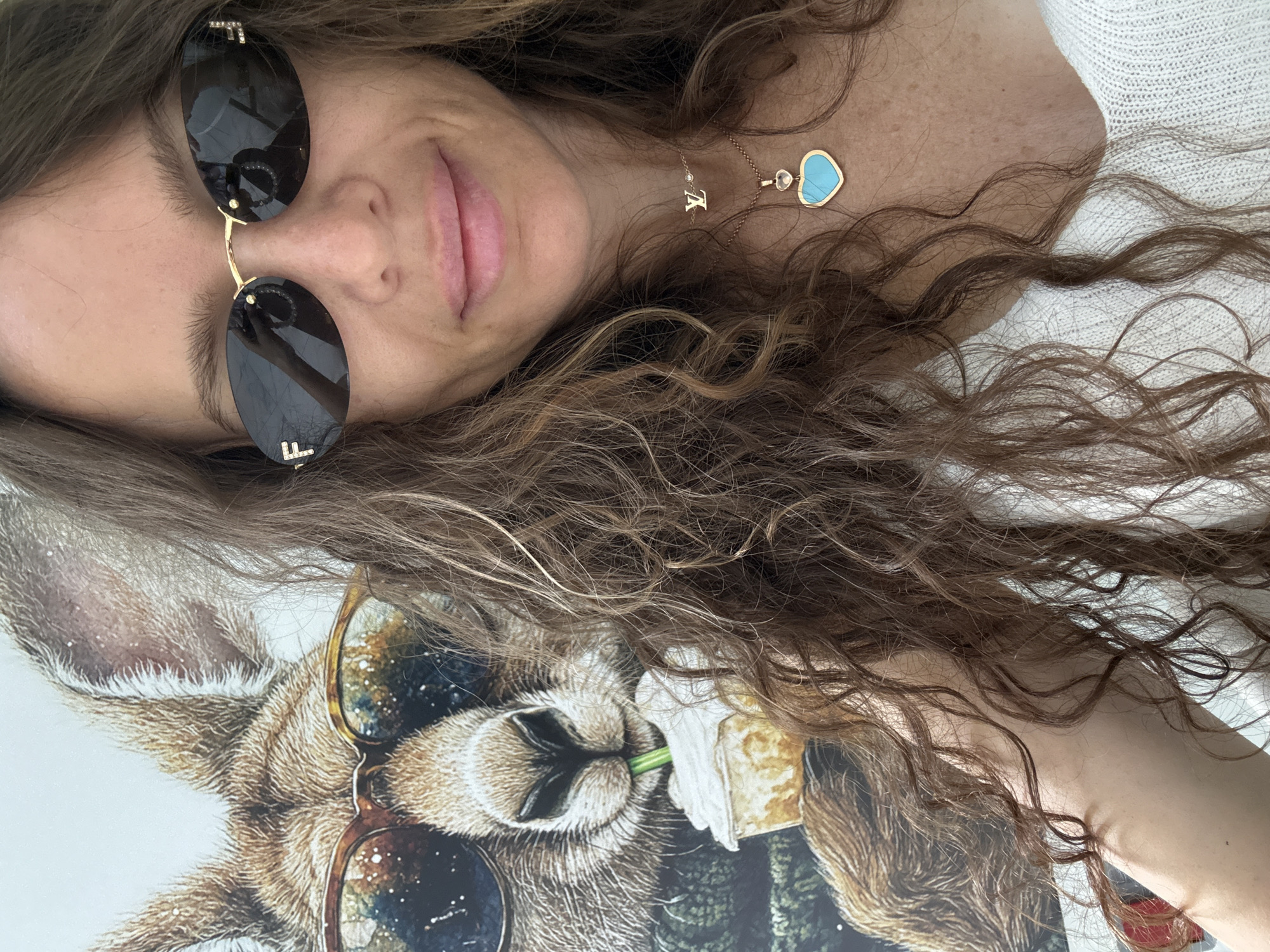 An adult female with long, wavy brown hair is taking a selfie. She is wearing sunglasses with gold frames and what appear to be a gold letter "F" and a diamond on each side. She is also wearing two necklaces, one with a gold letter "V" and a small diamond, and another with a gold heart pendant containing a blue stone. She is wearing a white, textured top. Behind her, on a wall, is a large portrait of a kangaroo wearing sunglasses and drinking a beverage with a straw. The environment appears to be indoors, possibly in a home or cafe, and the lighting suggests daytime. There is no visible indication that this image was taken in Moscow, Russia.