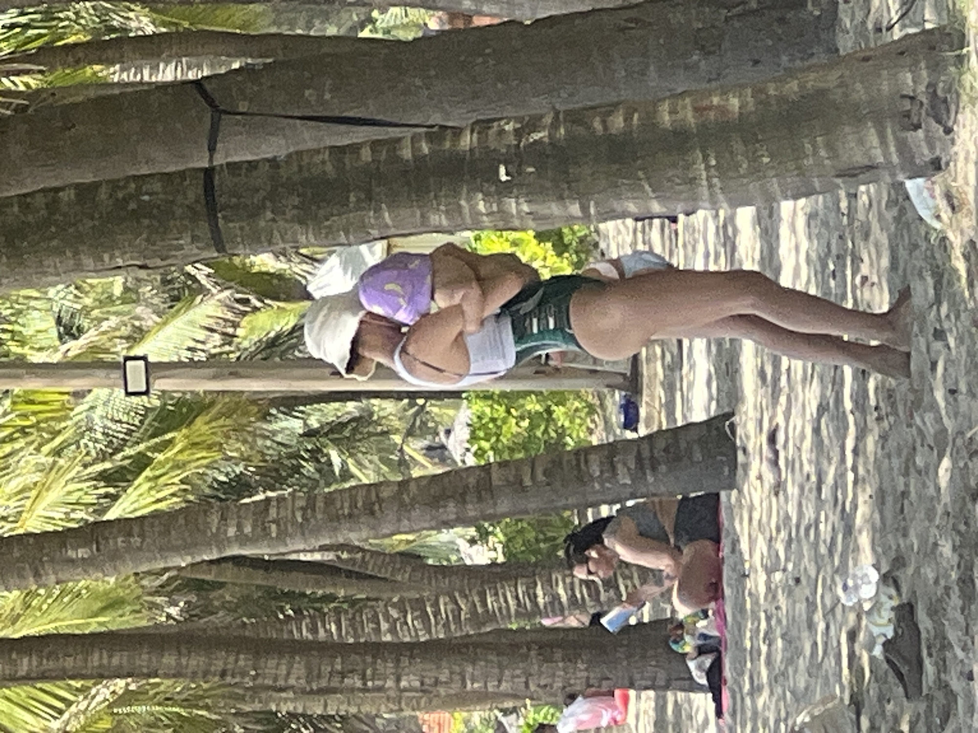 The image shows a beach scene, likely in Đà Nẵng, Vietnam, with several palm trees casting shadows on the sandy shore. In the foreground, a woman in a white bucket hat and a green one-piece swimsuit is holding a child wearing a purple hat. The woman is facing away from the camera, and the child is being held in her arms.
Further back, another woman is sitting on a pink mat, reading a book. She is wearing a grey bikini top and dark shorts. Various beach items, including towels and a bag, are scattered around her. In the background, there are other beachgoers, some with umbrellas, and lush green vegetation. The overall scene depicts a casual day at the beach.